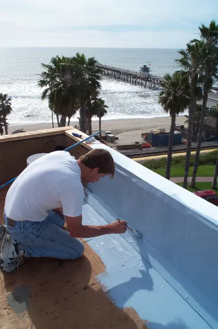 Dan, Cool Roof Company foreman and lay pastor, kneeling to detail the parapet waterproofing at the Sea Horse Inn oceanfront restoration in San Clemente, with the pier visible behind him