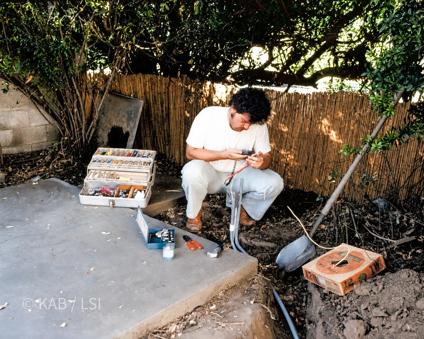 A worker crouched beside an open trench splicing electrical wire, with a toolbox, a shovel, and a box of TW-12 wire on the ground.