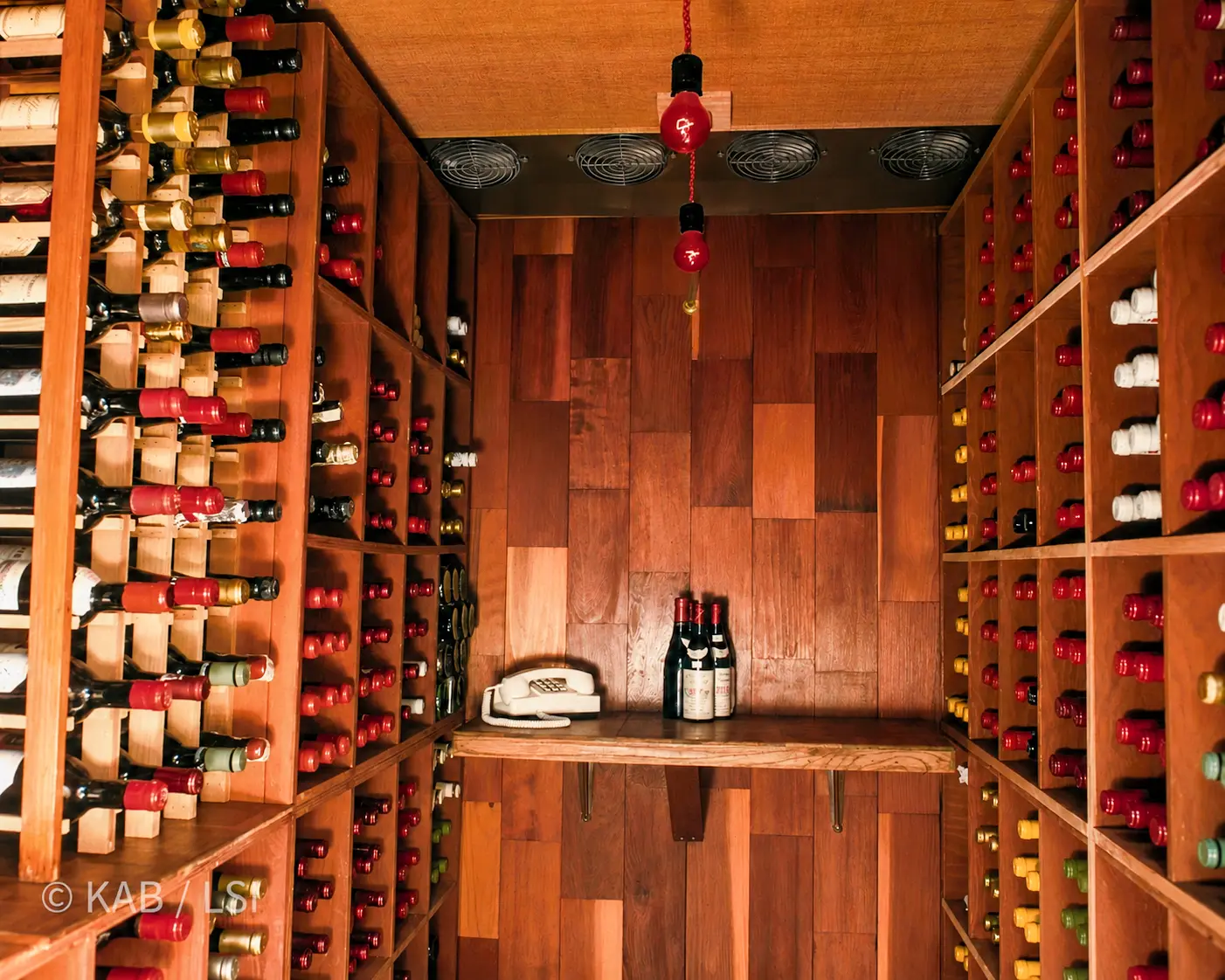 A finished wine cellar interior with full redwood racking, hundreds of bottles, a four-fan refrigeration array overhead, and a corded telephone on the uncorking shelf.