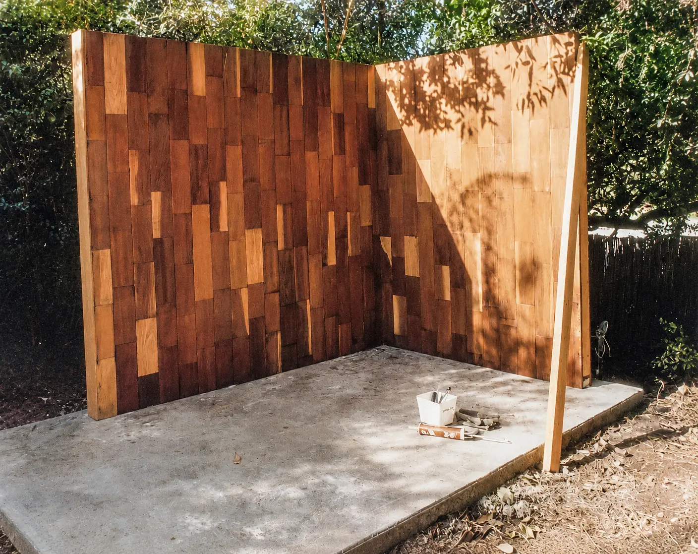 Two redwood-clad cellar walls standing on a freshly poured concrete pad, with a caulk gun and trowels on the slab.