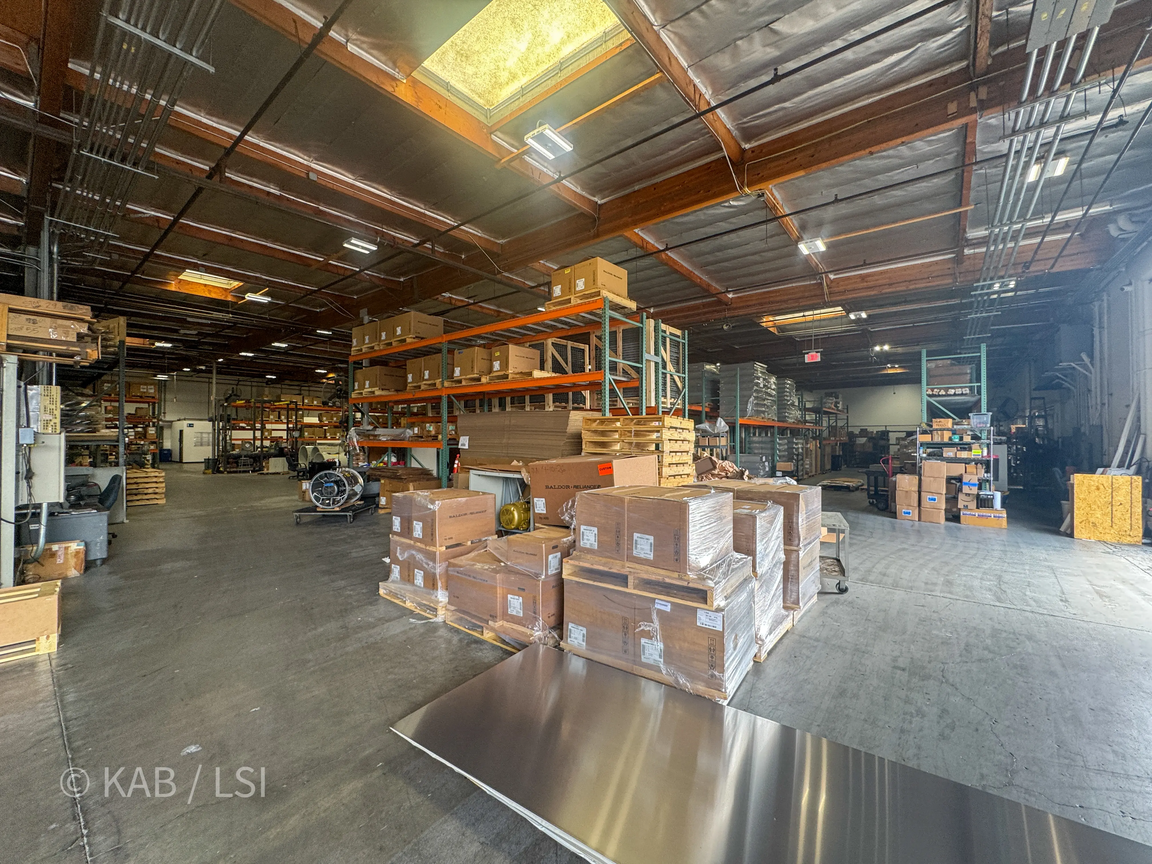Wide interior view from mid-building looking deeper into the ToroAire manufacturing floor, showing pallets of Baldor-Reliance motors, sheet metal stock, pallet racking, and a rooftop skylight overhead.