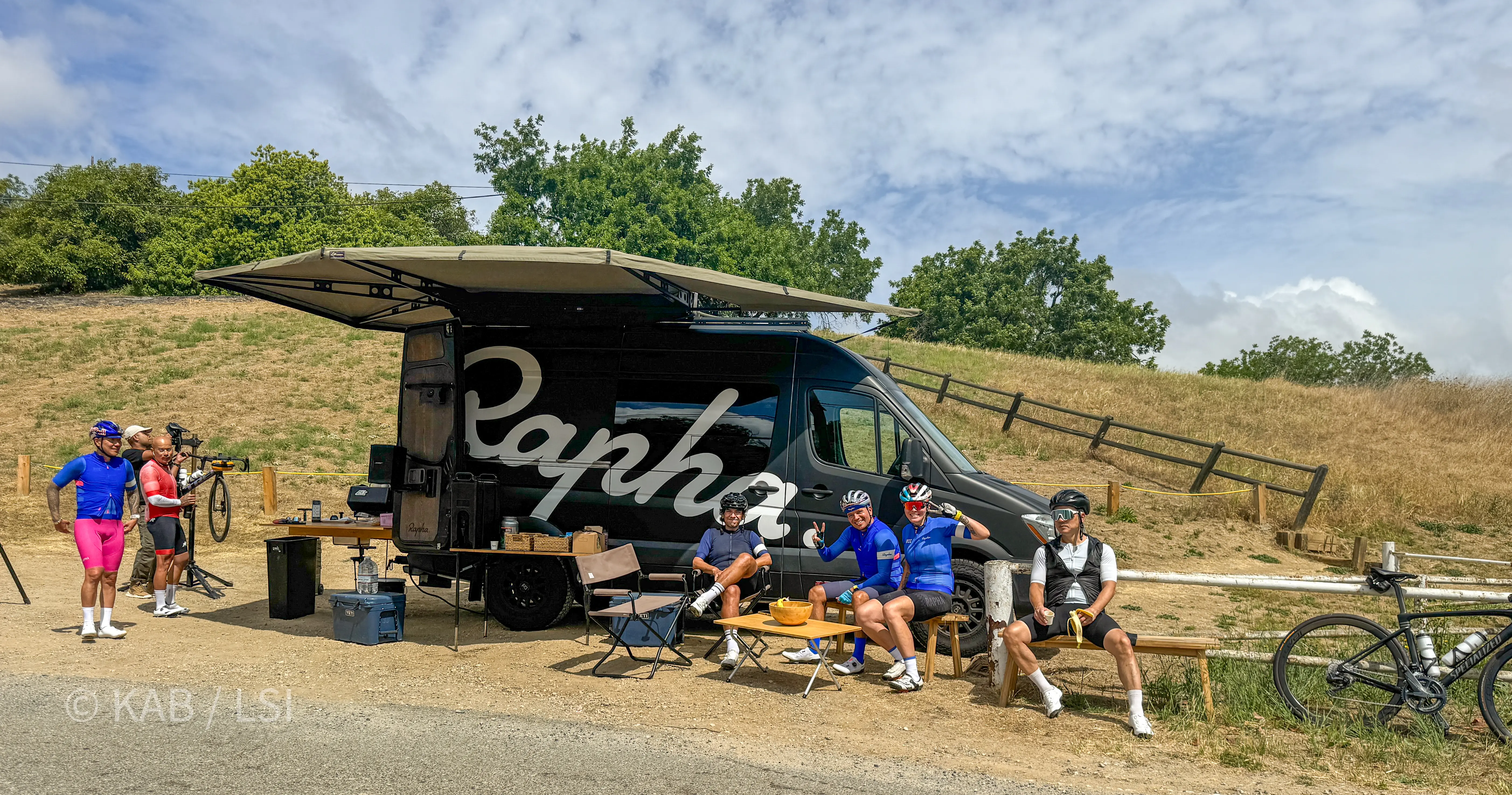 A Rapha cycling team support van set up on a La Habra Heights hillside roadside, awning extended over a folding table where weekend riders rest with bikes leaning against a wooden fence