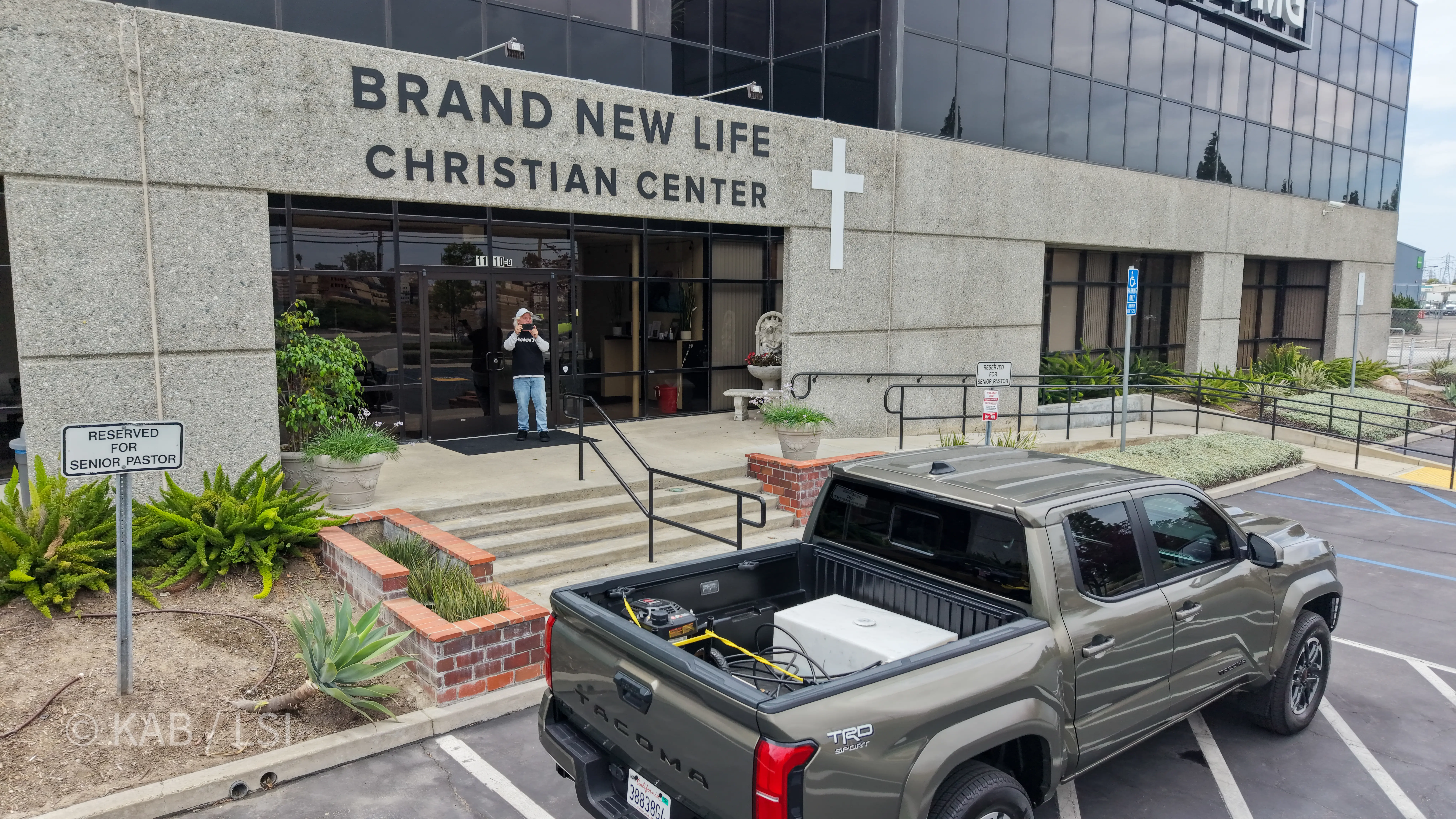 Front entrance of Brand New Life Christian Center at 1110 W. Taft Avenue, Orange, California, with the church's full name and cross on the building's stone facade. Longtime church volunteer Jeff Billig stands at the doorway in the foreground after locking up, photographing the drone overhead. A Reserved for Senior Pastor parking sign and ornamental aloe planter in the foreground.