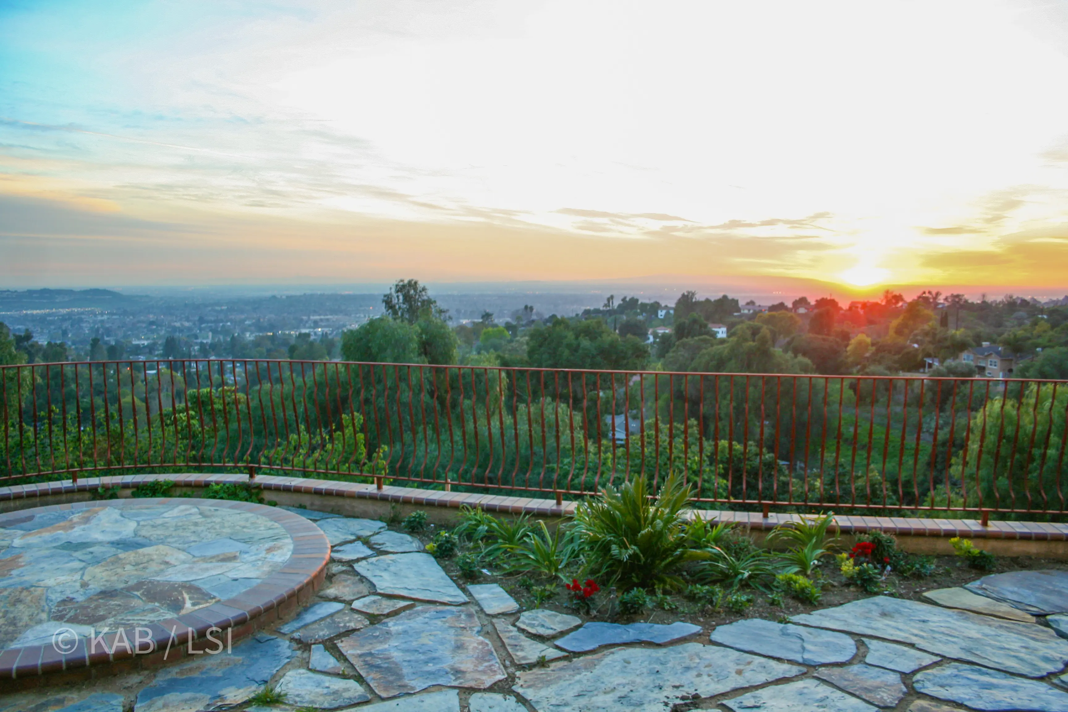 Sunset from an observation deck above La Habra Heights — wrought-iron railing, brick-and-flagstone patio, city falling away in the valley below.
