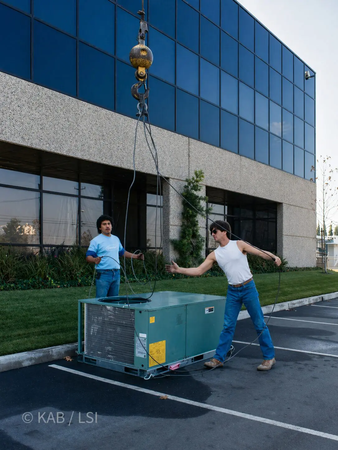 1987. Keith Bennett at 34, in a white tank, sunglasses, blue jeans and work boots, leaning back on a tag line as a five-ton Carrier package air conditioning unit is rigged onto its roof curb at the MDB Systems 88,000 sq ft building at 1110 W. Taft Avenue, Orange, California. A second worker in a light blue tank holds the opposite tag line. The dark blue mirror-glass curtain wall and stone band of the new building are already in place behind them.