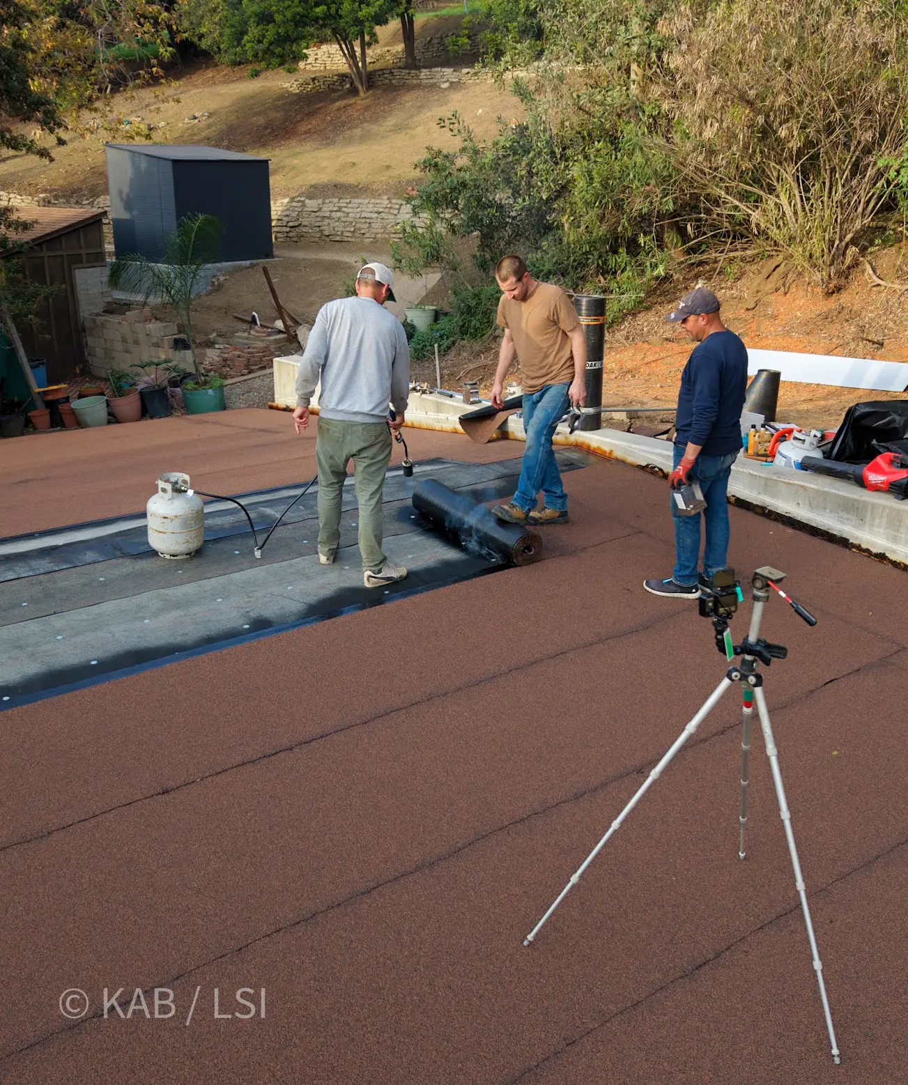 GoPro on tripod in foreground, three-man torch-down roofing crew working on a neighbor’s horse barn in the background
