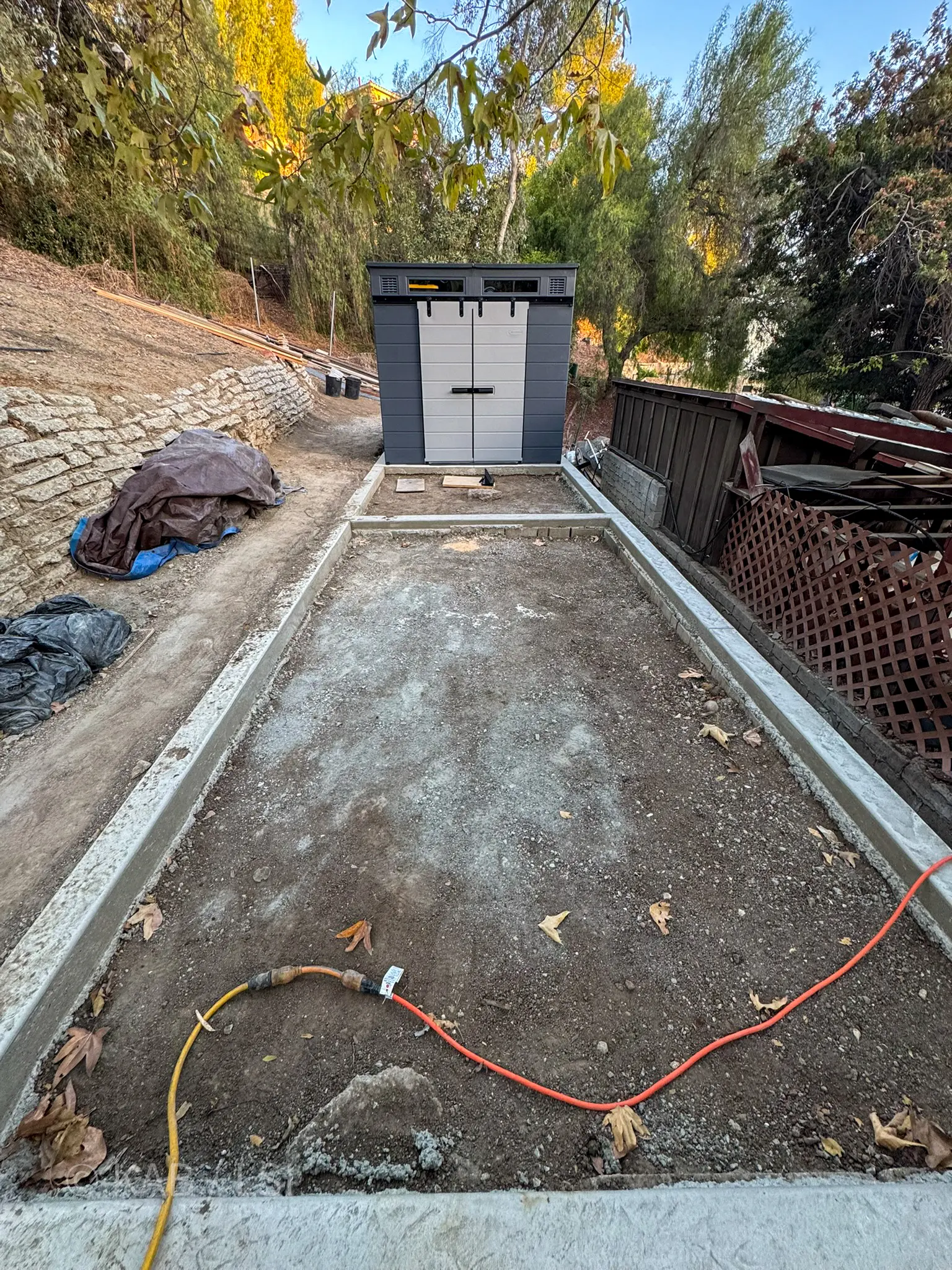 Reinforced concrete curb framing a second-story tier on a La Habra Heights hillside property, prepared for pavers and artificial grass, with a Suncast polymer shed installed at the far end