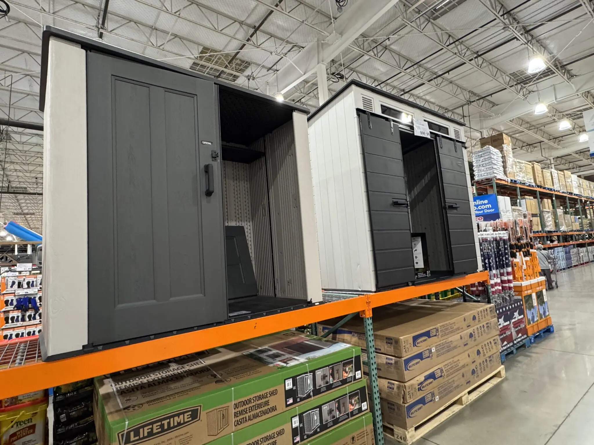 Two polymer outdoor storage sheds displayed on an orange Costco warehouse rack, with Lifetime and Suncast flat-pack cardboard boxes stacked on pallets below — the aisle every La Habra Heights homeowner has walked past