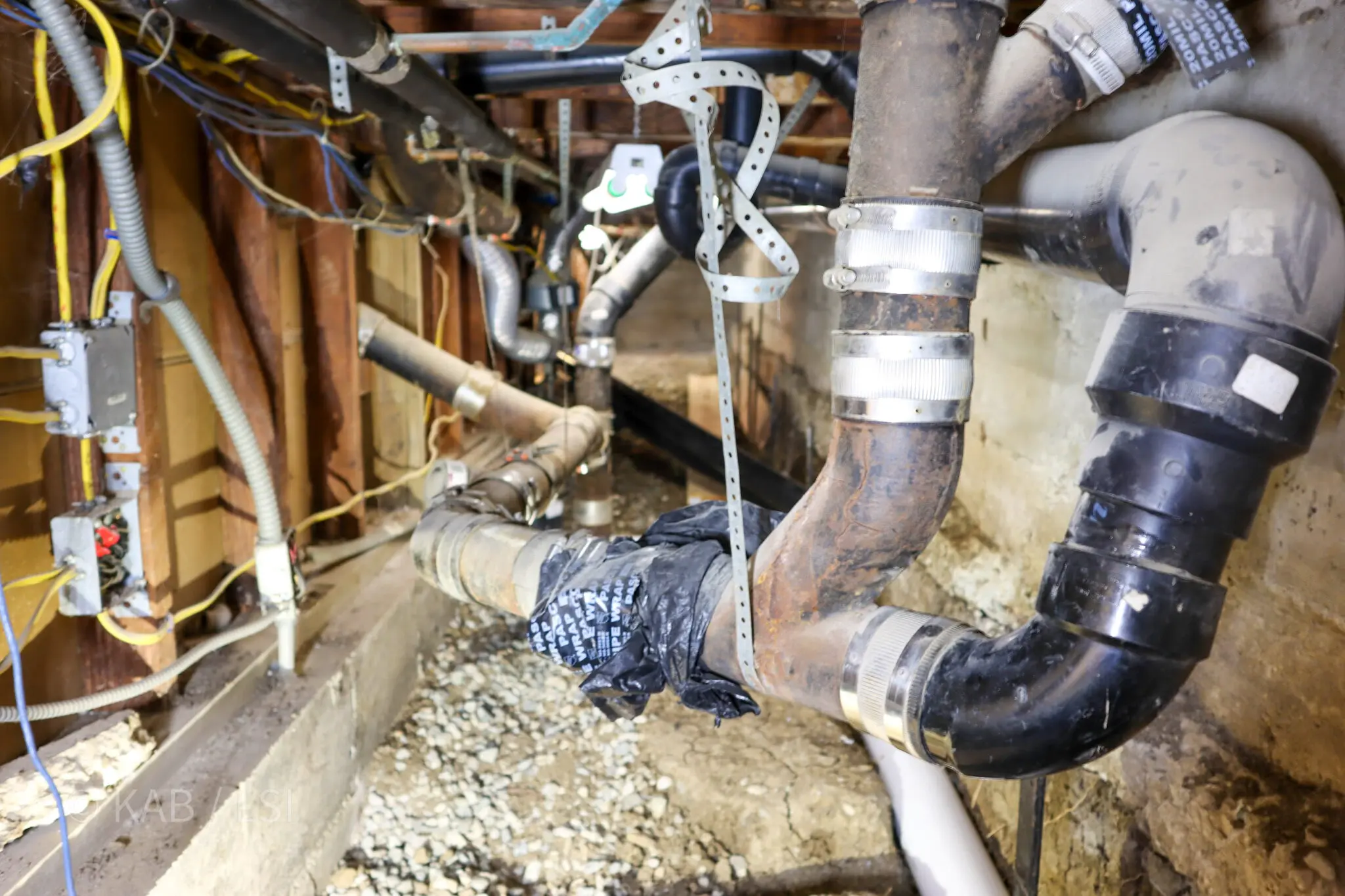 Crawlspace beneath a hillside home showing corroded cast iron drain lines patched with plastic couplings, band clamps and pipe wrap tape