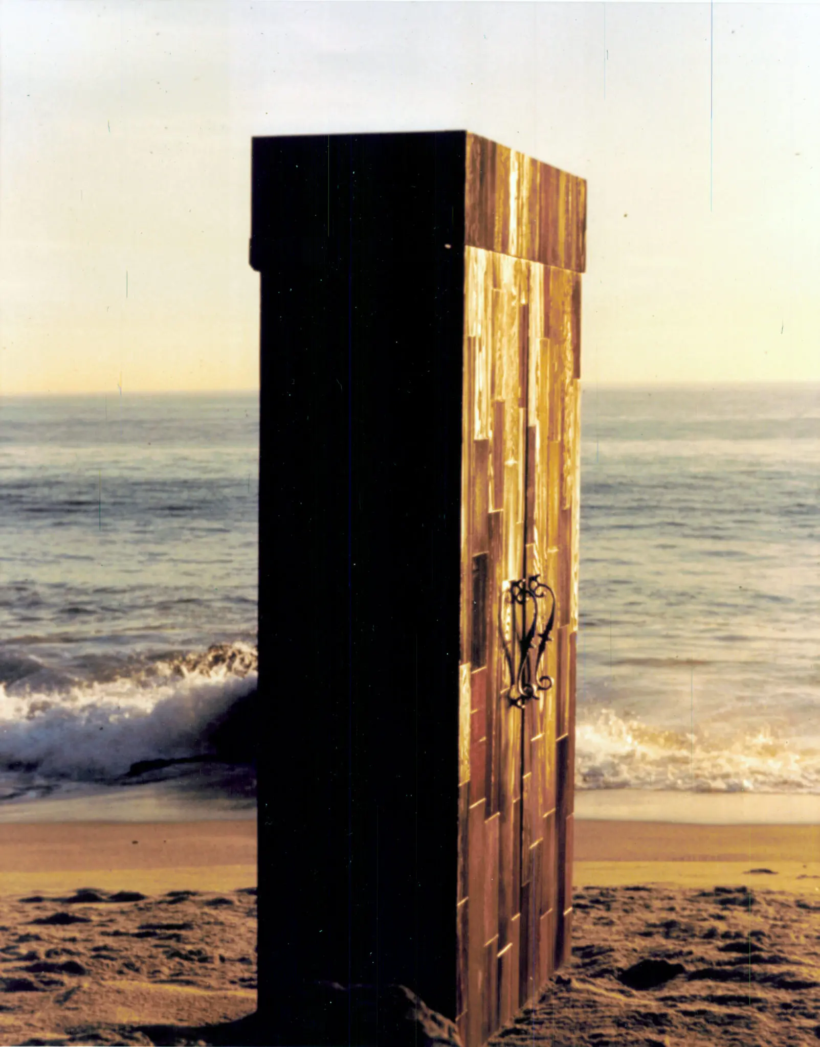 A redwood-clad free-standing wine cellar cabinet with a wrought-iron handle, standing upright on a Malibu beach with the surf behind it.