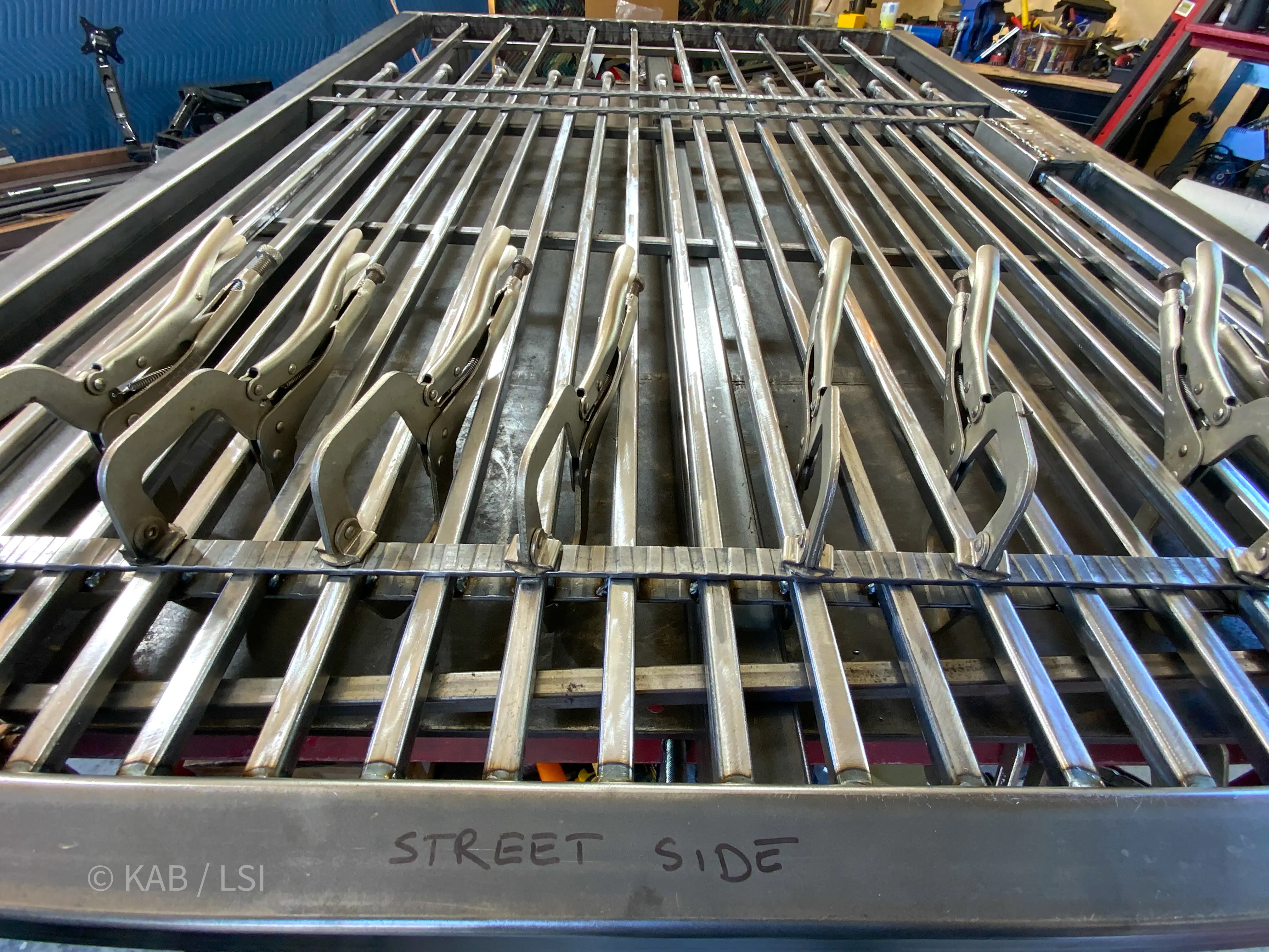 A coyote-proof wrought-iron fence panel under construction in a welding shop — lower pickets held to two inches on center, clamped for welding.