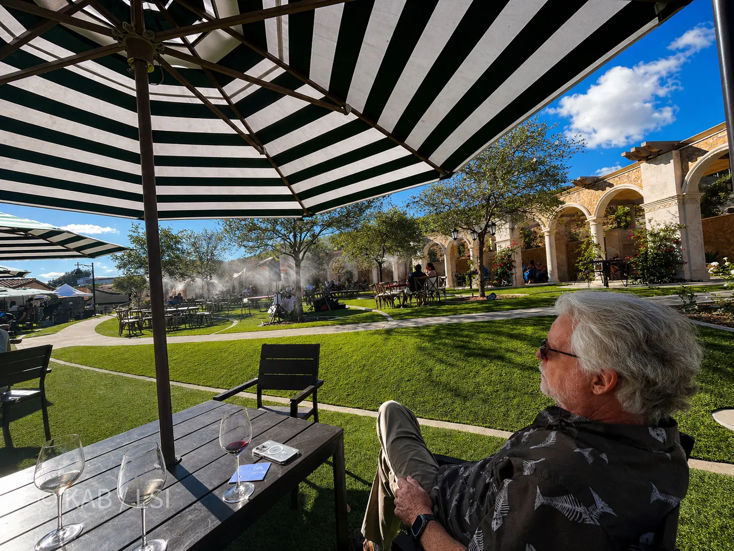 Keith Bennett seated alone at a slatted wood table under a green-and-white striped umbrella at Vienza, one of the village destinations at Europa Village winery in Temecula — taking in the lawn, the misters in the trees, the Romanesque arched colonnade, and the clusters of guests laughing in their own private spaces beyond.