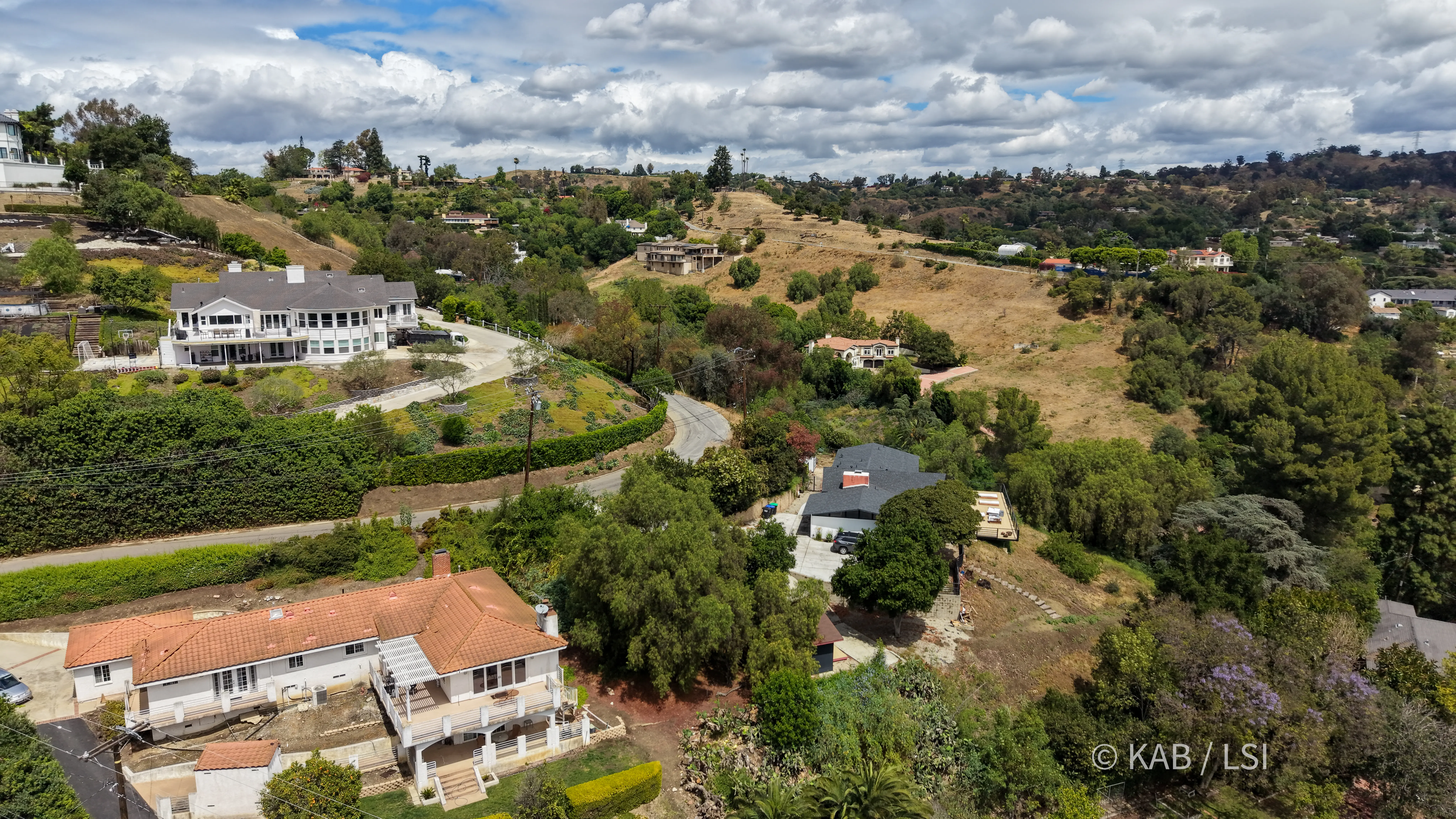 La Habra Heights hillside neighborhood in May 2026 — homes on one-acre-minimum parcels along a winding hillside road, with irrigated greenery against early-summer chaparral.