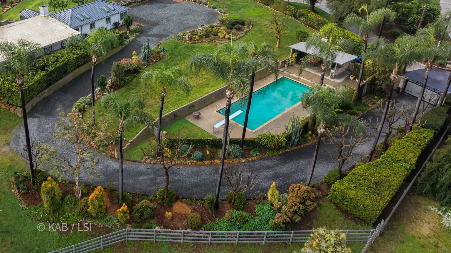 A private hillside pool terrace ringed by tall palms, with a guest house, looping gravel drives, and layered garden beds, observed during a drone flyover on the east side of La Habra Heights.