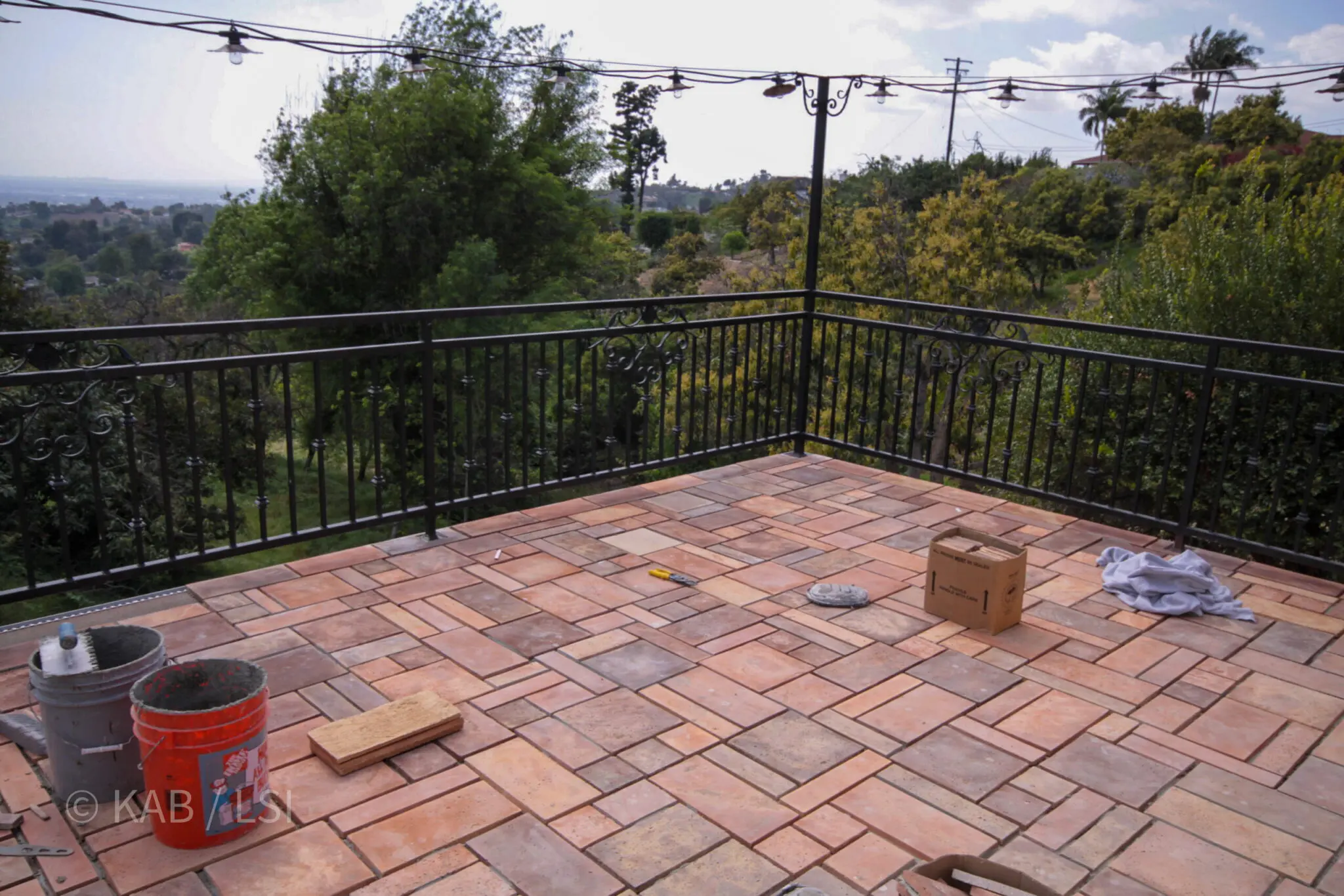 A wrought-iron-railed terracotta tile deck under construction off an upstairs master suite on a La Habra Heights hillside — string lights overhead, the valley and treetops beyond.