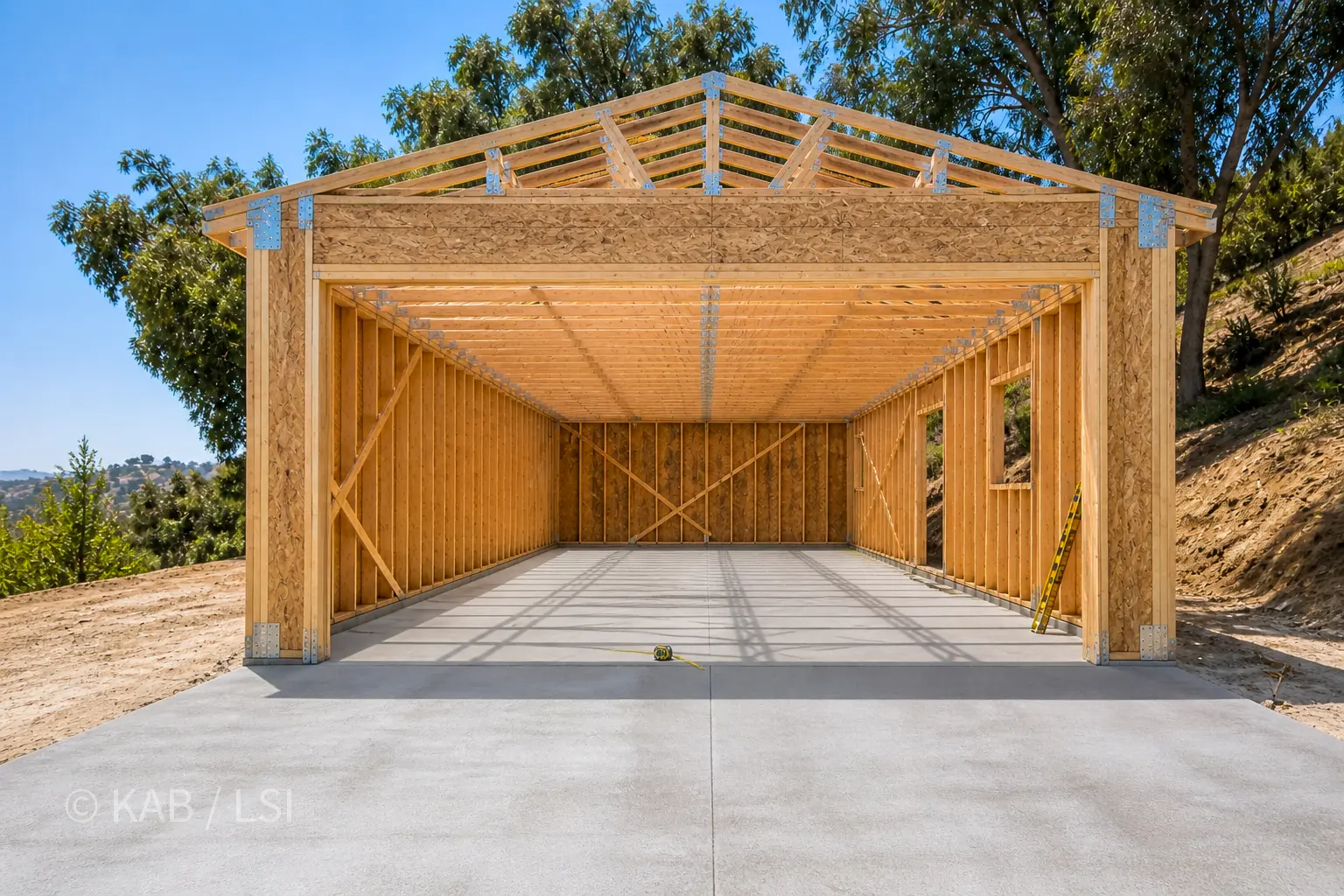A custom RV garage on a hillside lot mid-construction — freshly poured 20-by-60 reinforced concrete slab, engineered roof trusses spanning the full depth, OSB-sheathed walls with Simpson Strong-Wall corner hardware, tall front opening sized for a Class A motor home.