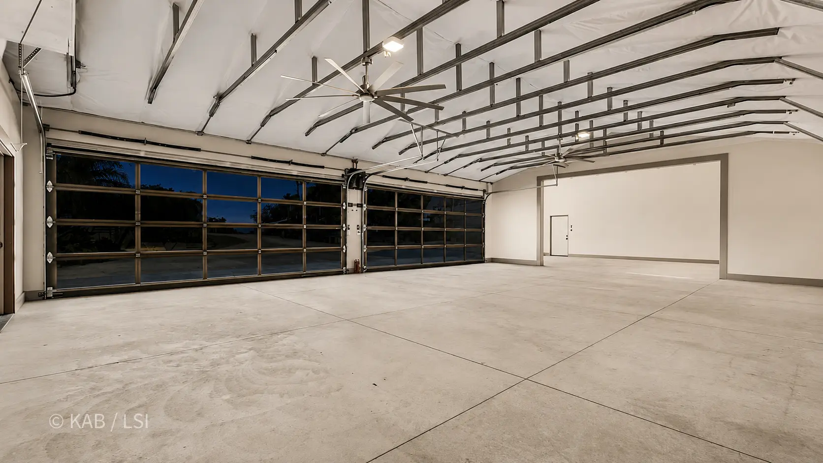 The empty interior of a hillside RV garage at dusk — exposed steel rafters, big-blade industrial fans, polished concrete floor.