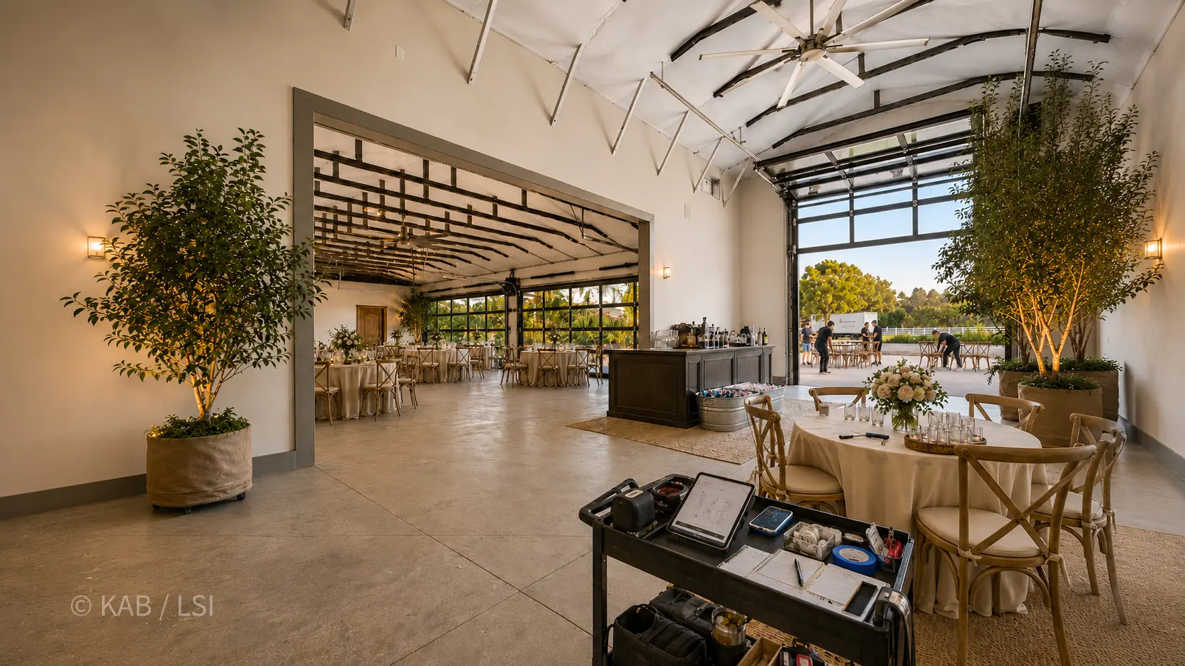 Inside an RV garage set for a wedding reception — round tables, cross-back chairs, bar service, glass garage doors rolled up to the patio.