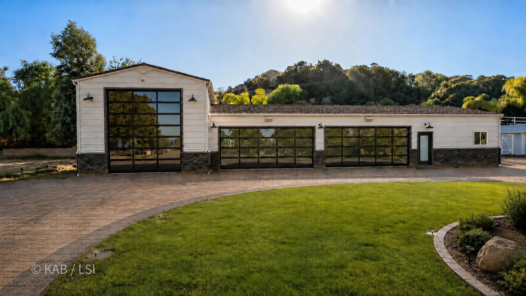 The exterior of a hillside RV garage at golden hour — full-height glass doors, white board-and-batten, paver apron.