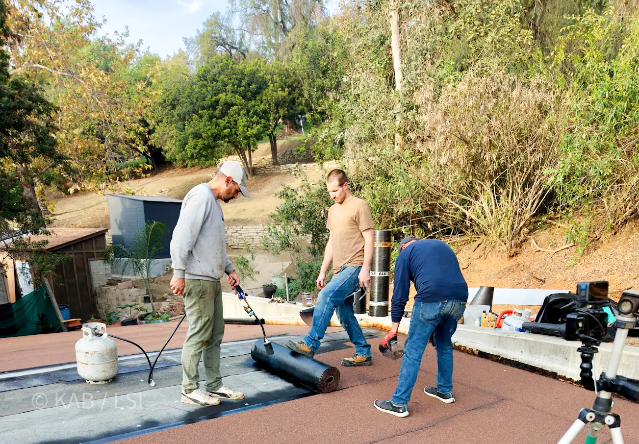 A La Habra Heights homeowner and his crew finish a torch-down roof on a hillside outbuilding before the storm arrives, viewed from above.