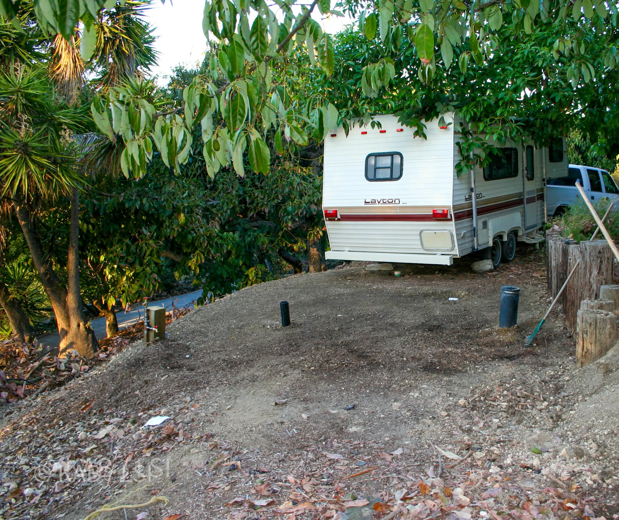 Layton fifth-wheel trailer parked beside a Water + Power Station under an avocado tree on a La Habra Heights hillside property during a whole-house makeover, serving as temporary quarters for the homeowner's live-in chef and housekeeper while the guest room was under construction — the water station visible at lower left, installed months earlier for wildfire defense, here repurposed with a pressure regulator stepping 125 PSI system pressure down for the trailer