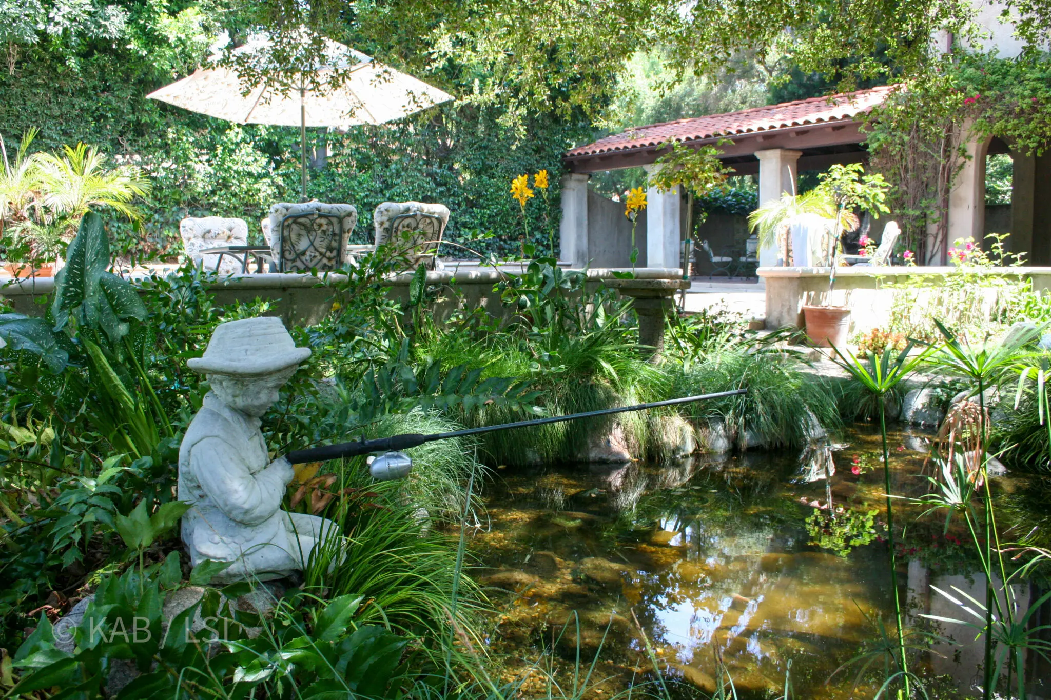 Stone statue of a boy fishing at the edge of a koi pond in a lush hillside garden; cushioned patio chairs under a parasol and a tile-roofed loggia in the background.