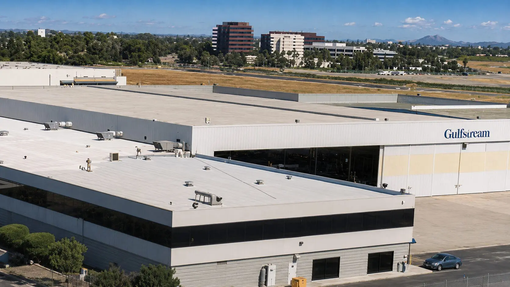 Aerial view of the Gulfstream facility at Long Beach Airport, a roofing crew spraying white cool-roof coating across the corporate-offices roof in the foreground, with the long hangar and a runway beyond.
