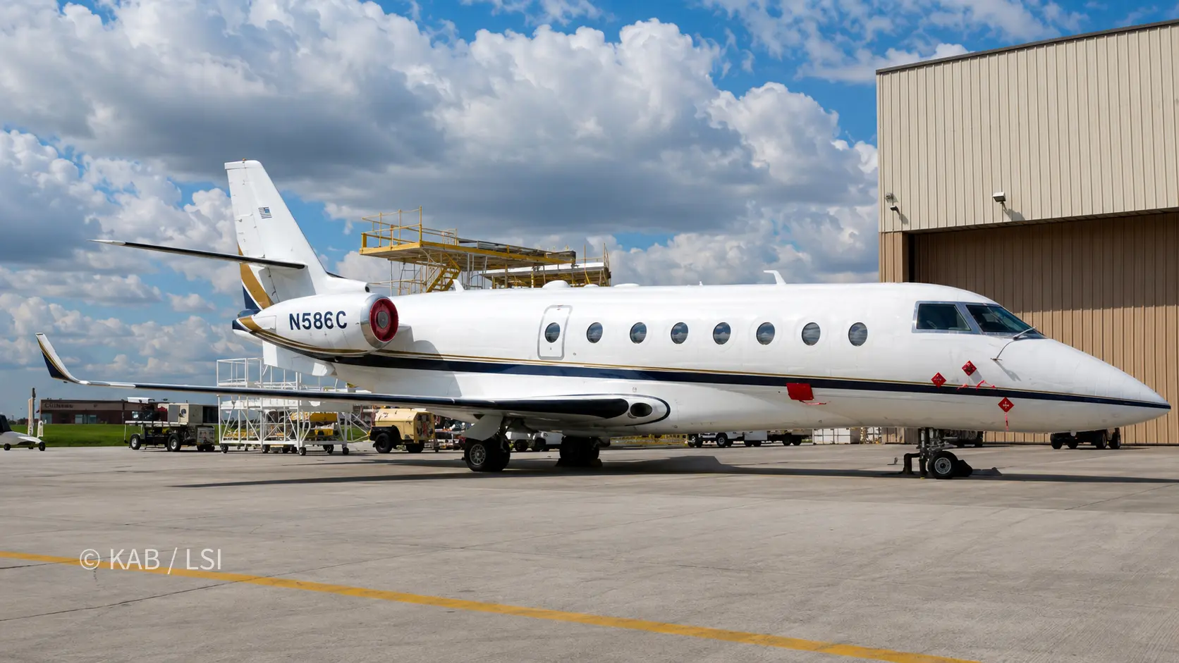 A large-cabin Gulfstream business jet parked beside a hangar at Long Beach Airport, maintenance scaffolding alongside, under a tall cloudy sky.