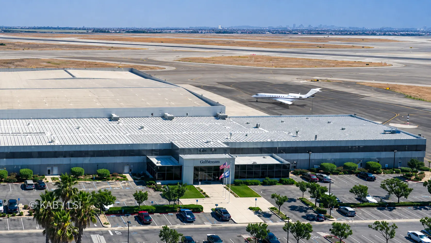 Aerial view of the Gulfstream facility at Long Beach Airport, its signed main entrance and flags in the foreground, a business jet on the airfield beyond, the Los Angeles basin stretching to the coast in the distance.