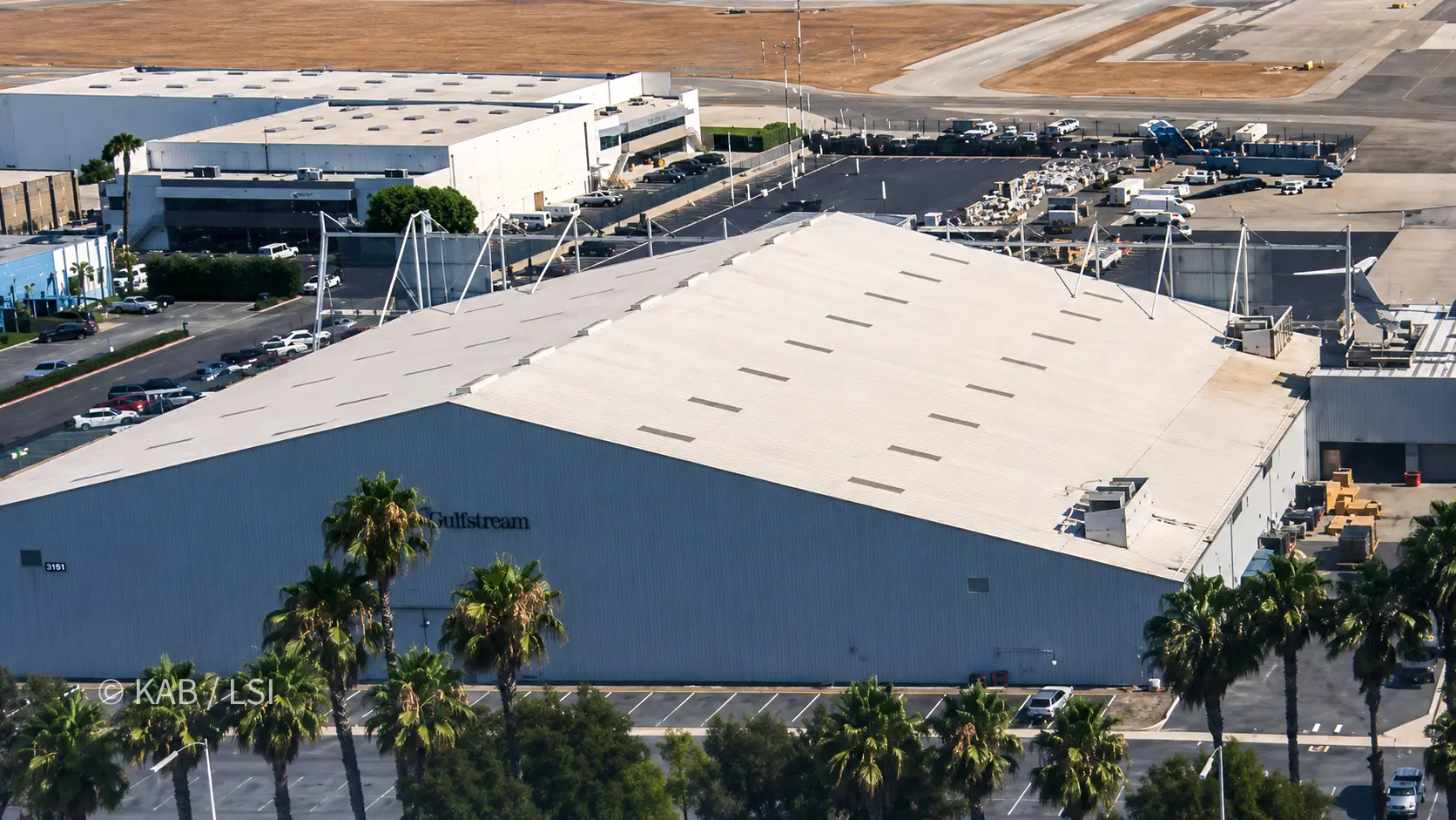 Aerial view of a large Gulfstream service hangar at Long Beach Airport, its broad pale roof, the Gulfstream name on the wall, palms and parking below, the airfield beyond.