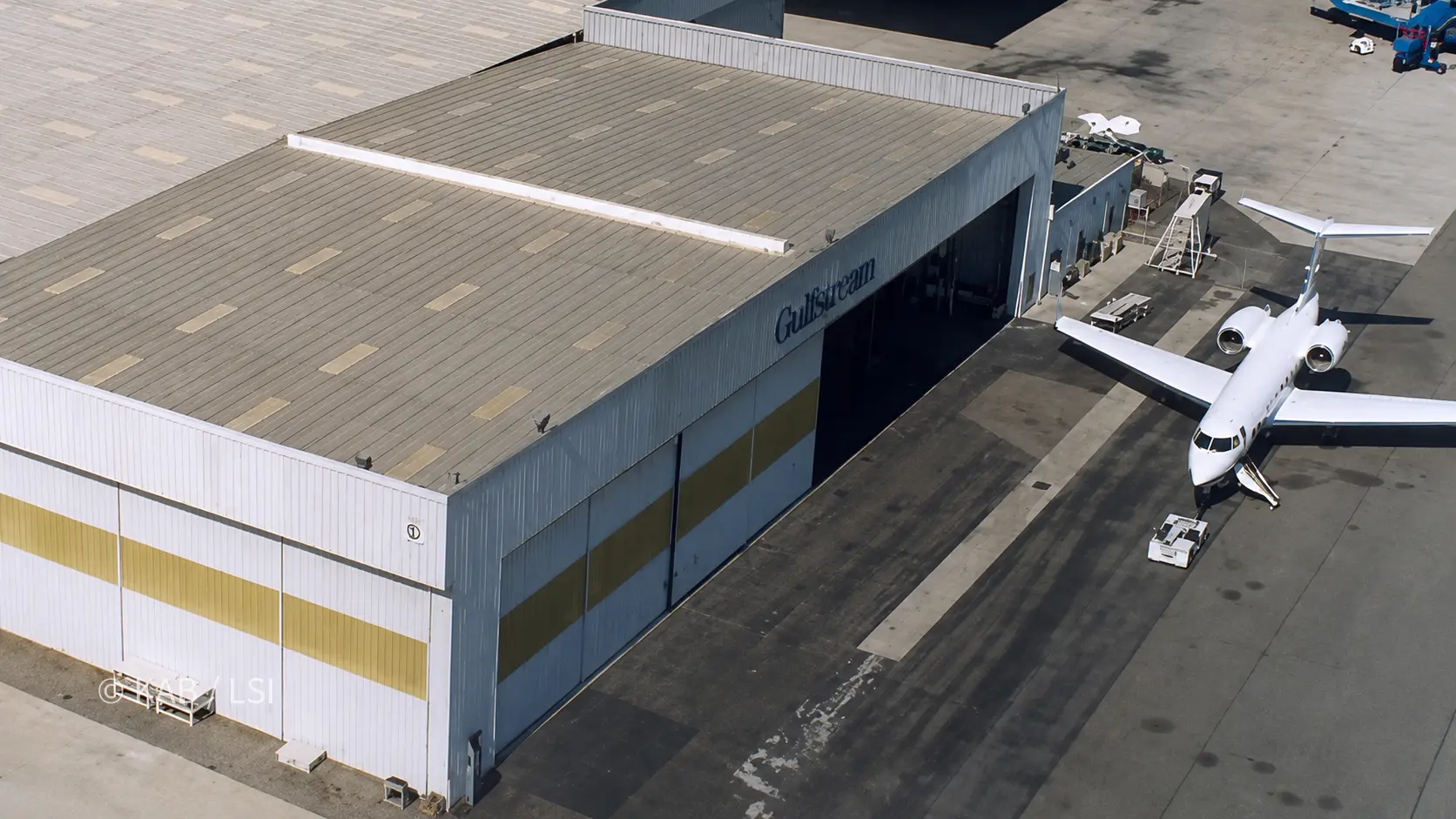 Aerial view of a Gulfstream hangar at Long Beach Airport, its tall doors open and a business jet on a tug at the entrance, the Gulfstream name across the wall.