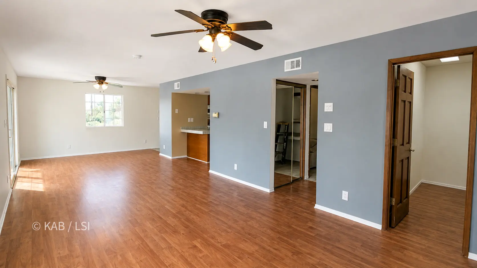 The ADU open great room from another angle: reddish hardwood floors, pale and blue-gray walls, two ceiling fans, a sliding glass door and window at left. At center, the kitchen pass-through and mirrored bifold closet doors; at right, an open wood door revealing the walk-in closet.