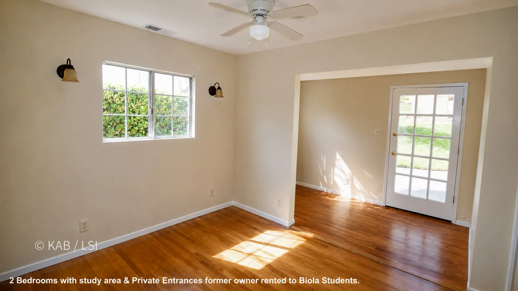 A bedroom with hardwood floors, beige walls, a ceiling fan, and two sconces flanking a window onto greenery. An opening at right leads to a light-filled study area with a glass-paned private-entrance door and a window looking onto a lawn.
