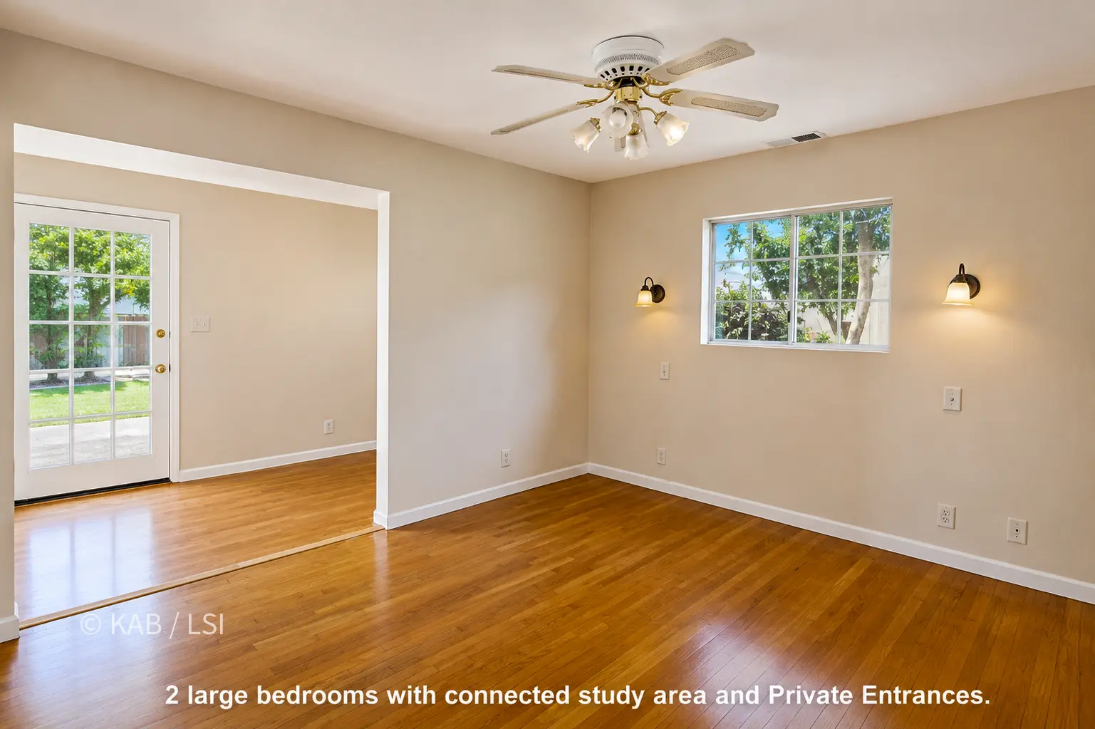 A bedroom with hardwood floors, beige walls, a ceiling fan, and two sconces flanking a window onto greenery. An opening at left leads to a study area with a glass-paned private-entrance door looking onto a lawn.