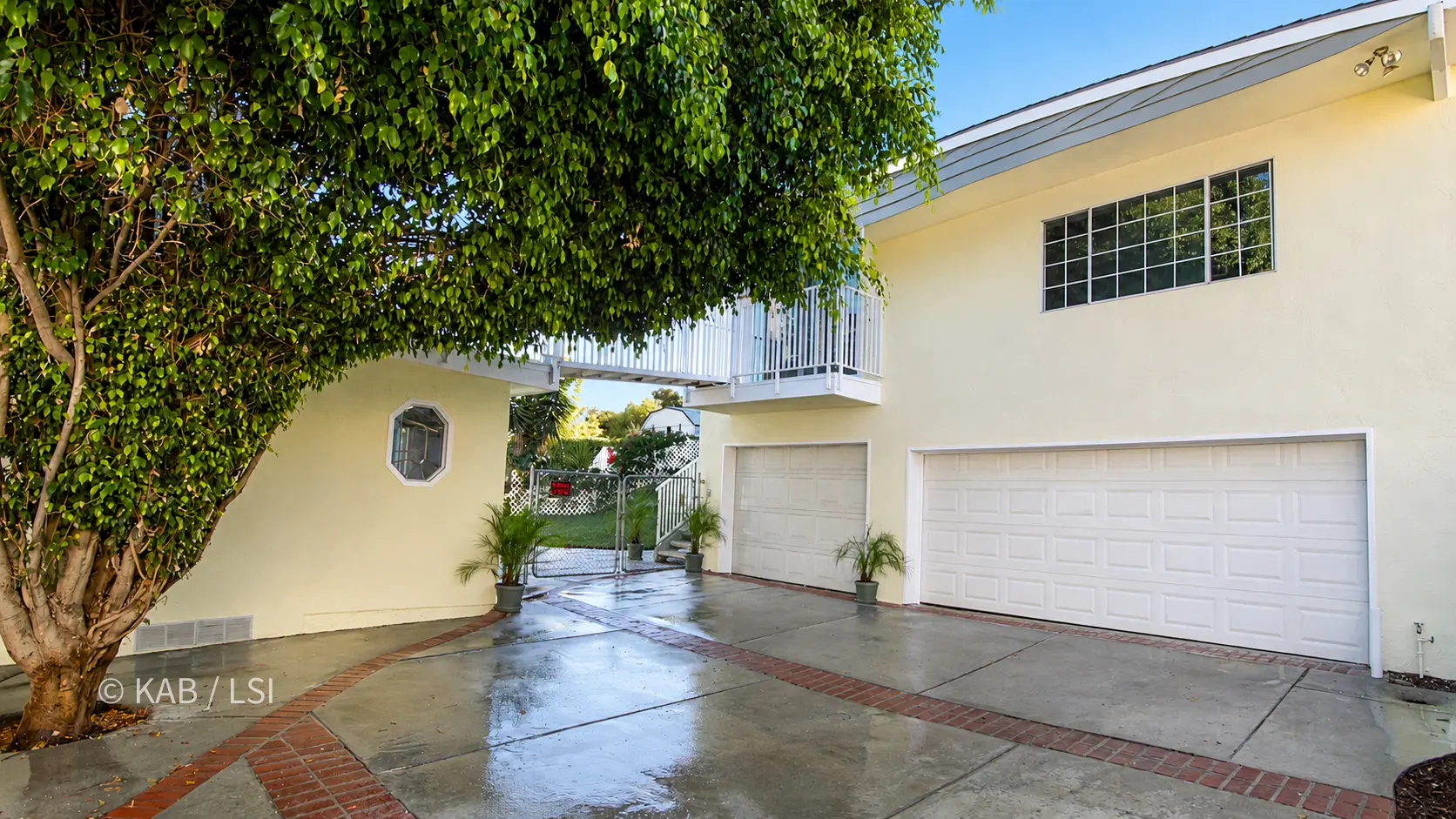 Wide view of a paved rear yard shaded by a large ficus tree. At left, a gable-roofed room with a sliding glass door; at center, the main house with a brick chimney, an octagonal window on a lower extension, and an elevated railed walkway to an upper deck; at right, the two-story building with the dwelling above the garage. Brick-banded concrete and potted palms throughout.