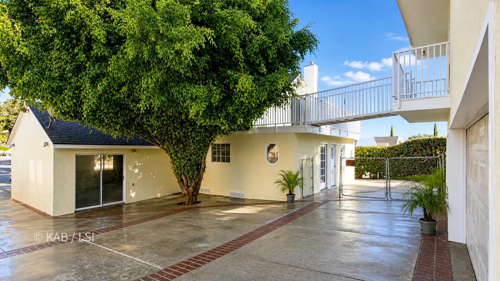Wide view of a brick-banded concrete rear yard shaded by a large ficus tree. At left, a gable-roofed room with a sliding glass door; at center, the main house with a brick chimney, an octagonal window, and French doors, with an elevated railed walkway and deck above; at right, the two-story building with a balcony and a garage door. Potted palms on the wet pavement.