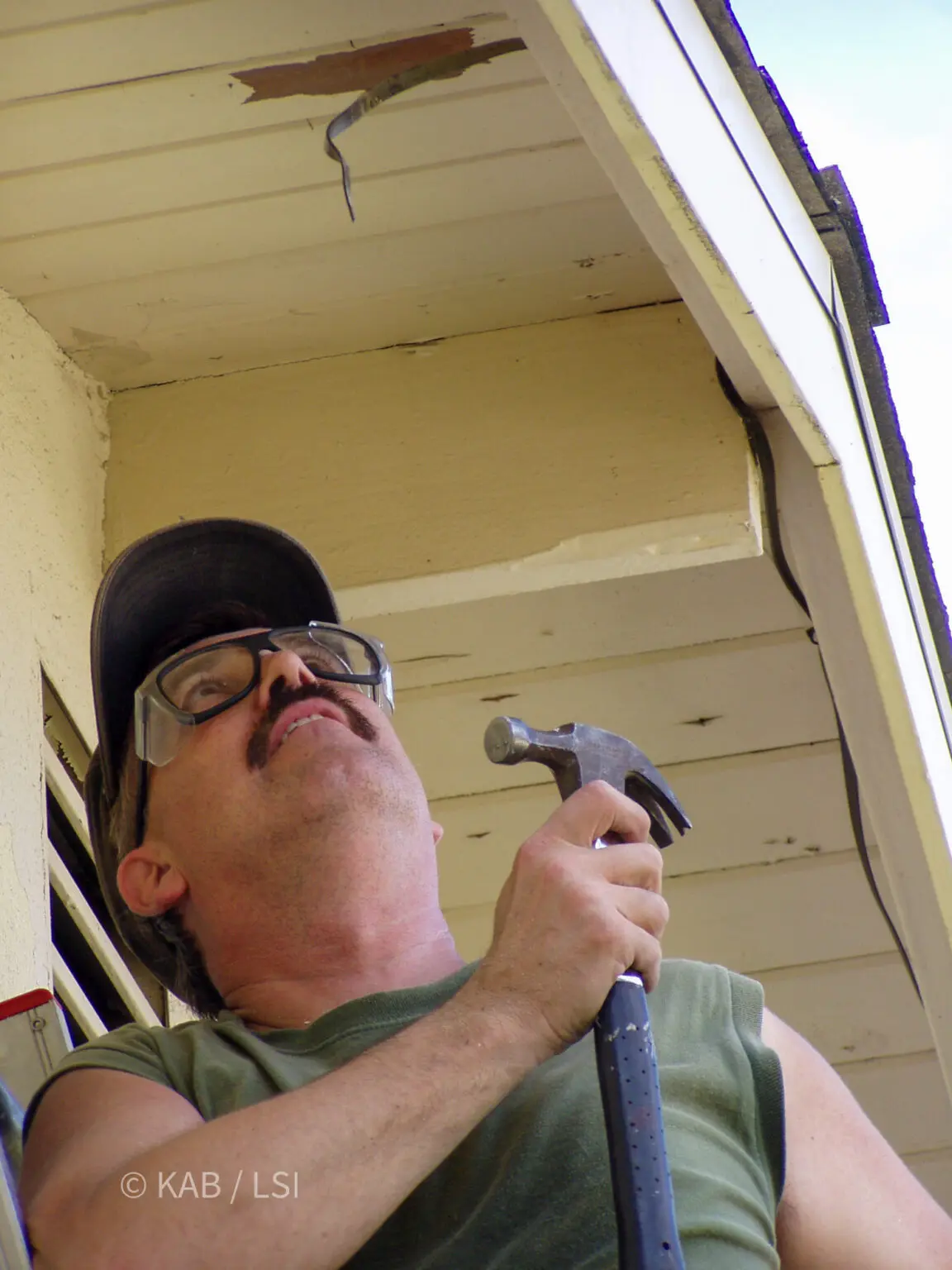 Carpenter looking up at a rotted soffit and fascia with a hammer raised, beginning demolition of a damaged roof overhang