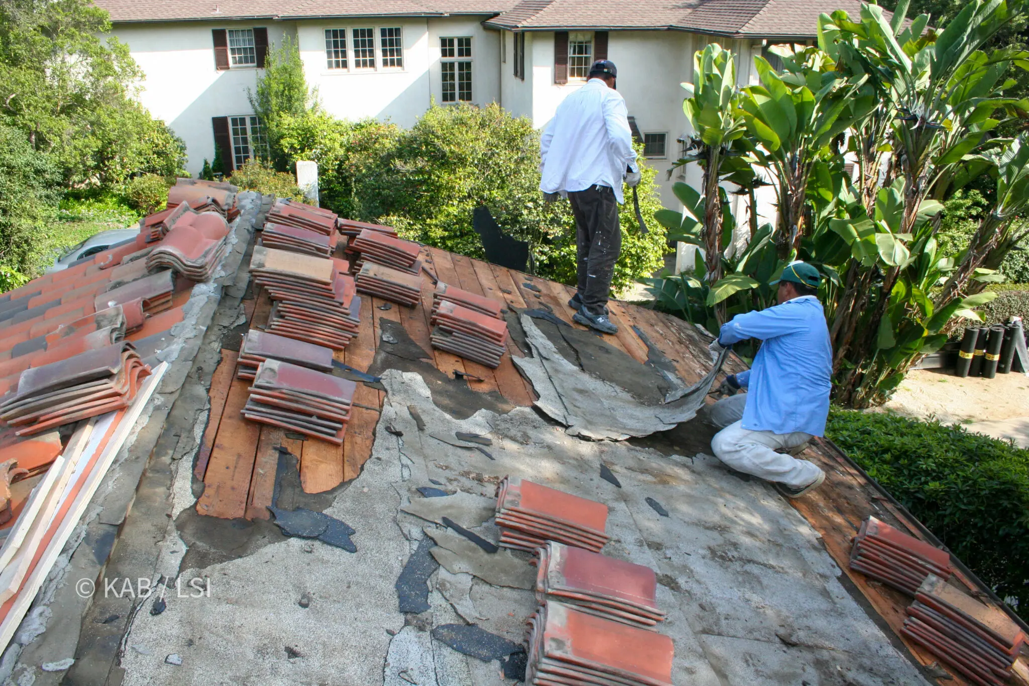 Roofers stripping failed underlayment from a 1920s clay tile roof with the original tile stacked in courses alongside