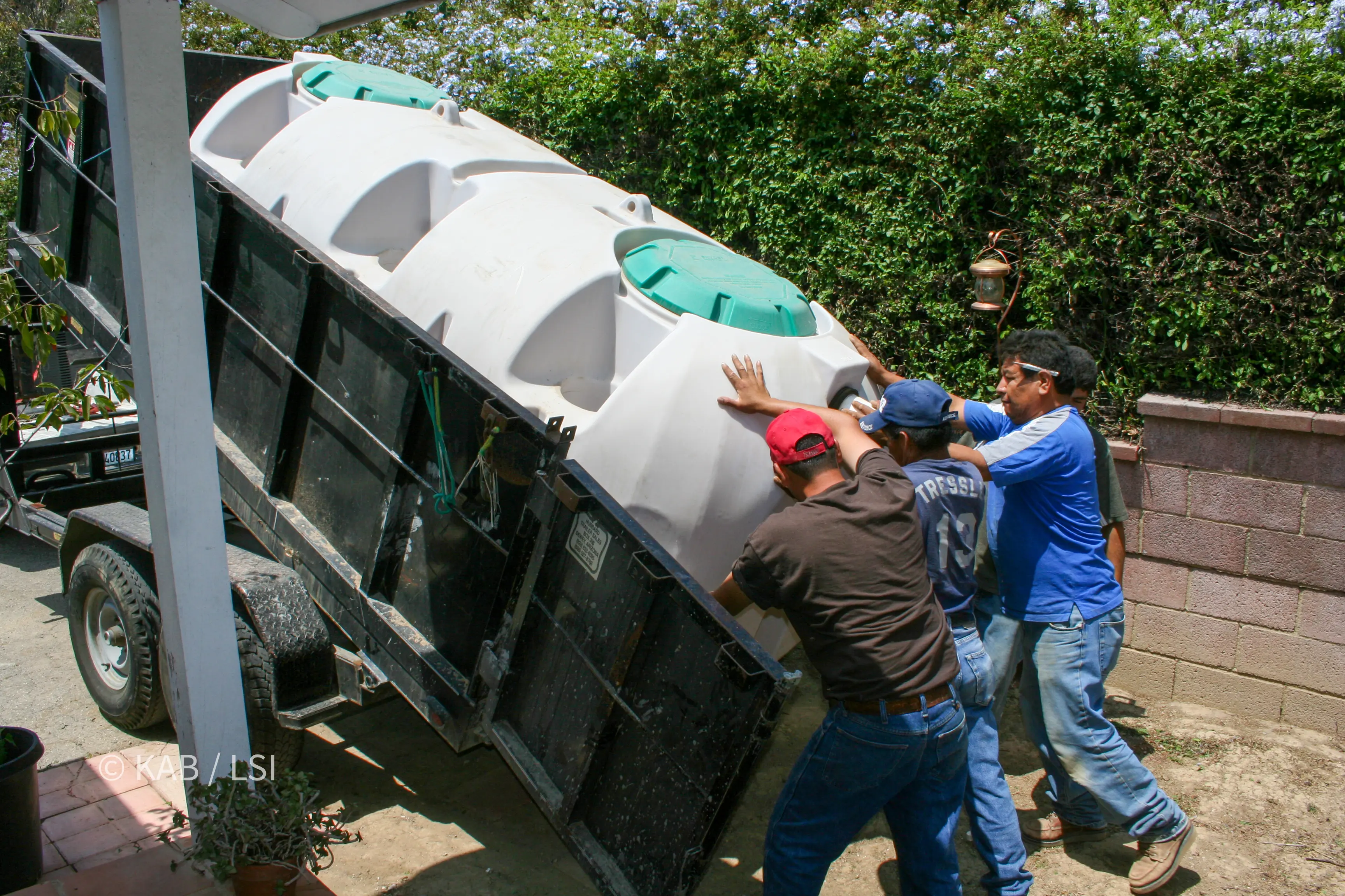 Four men guiding a 1,000-gallon polyethylene septic tank off a tipped trailer on a hillside driveway