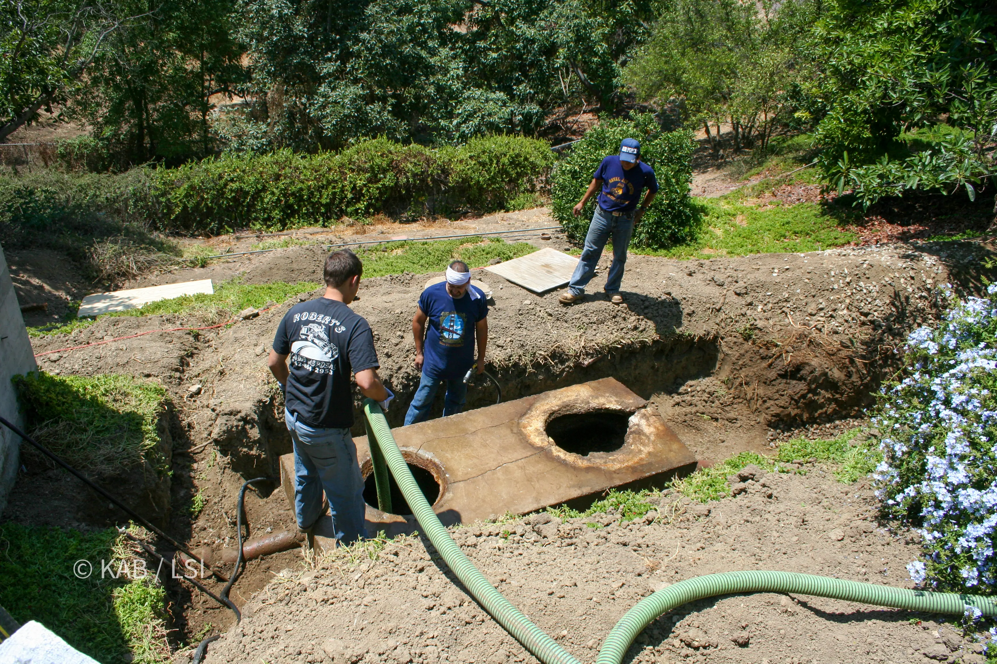Three workers at an excavated concrete septic tank with both chambers open, pumping and washing it before demolition