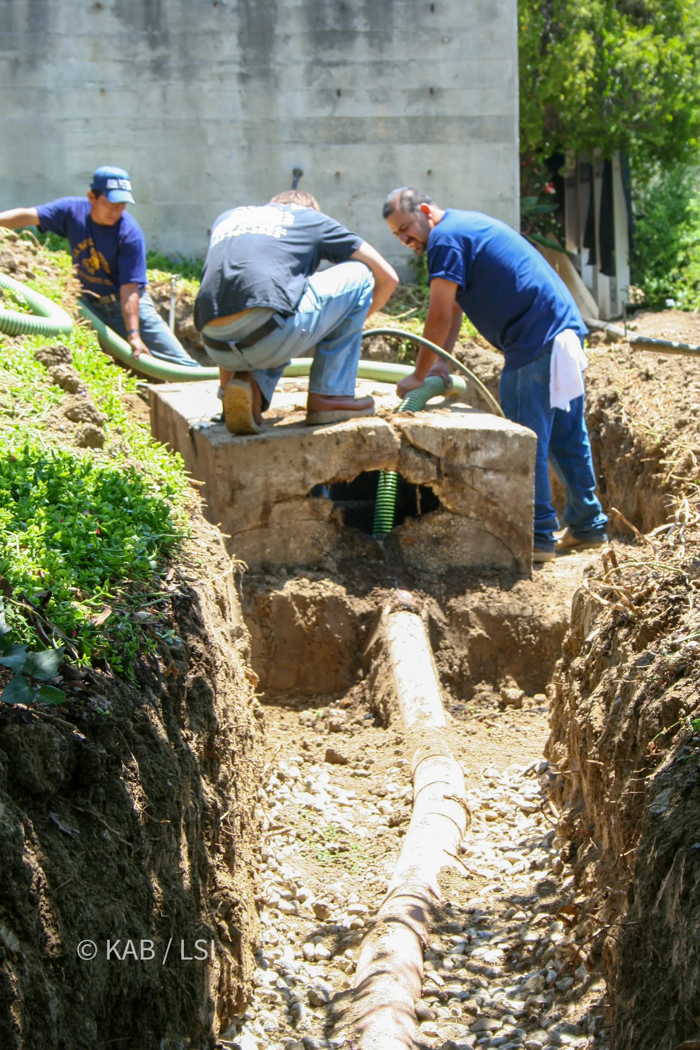 Narrow trench exposing an undersized 1950s clay leach line bedded in gravel beside a broken tank wall