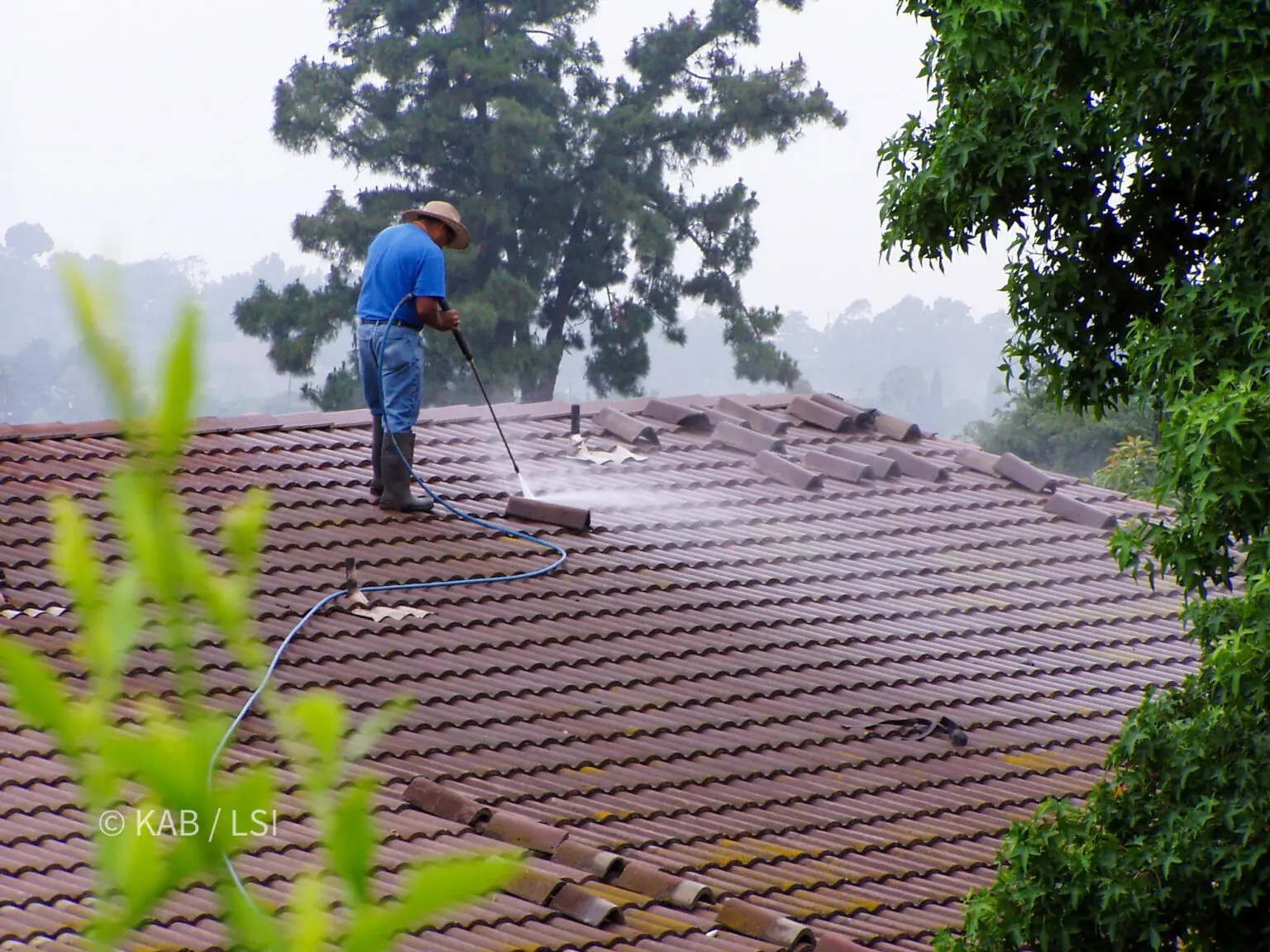 Roofer washing a concrete tile roof on an overcast hillside morning, tile stacked along the ridge