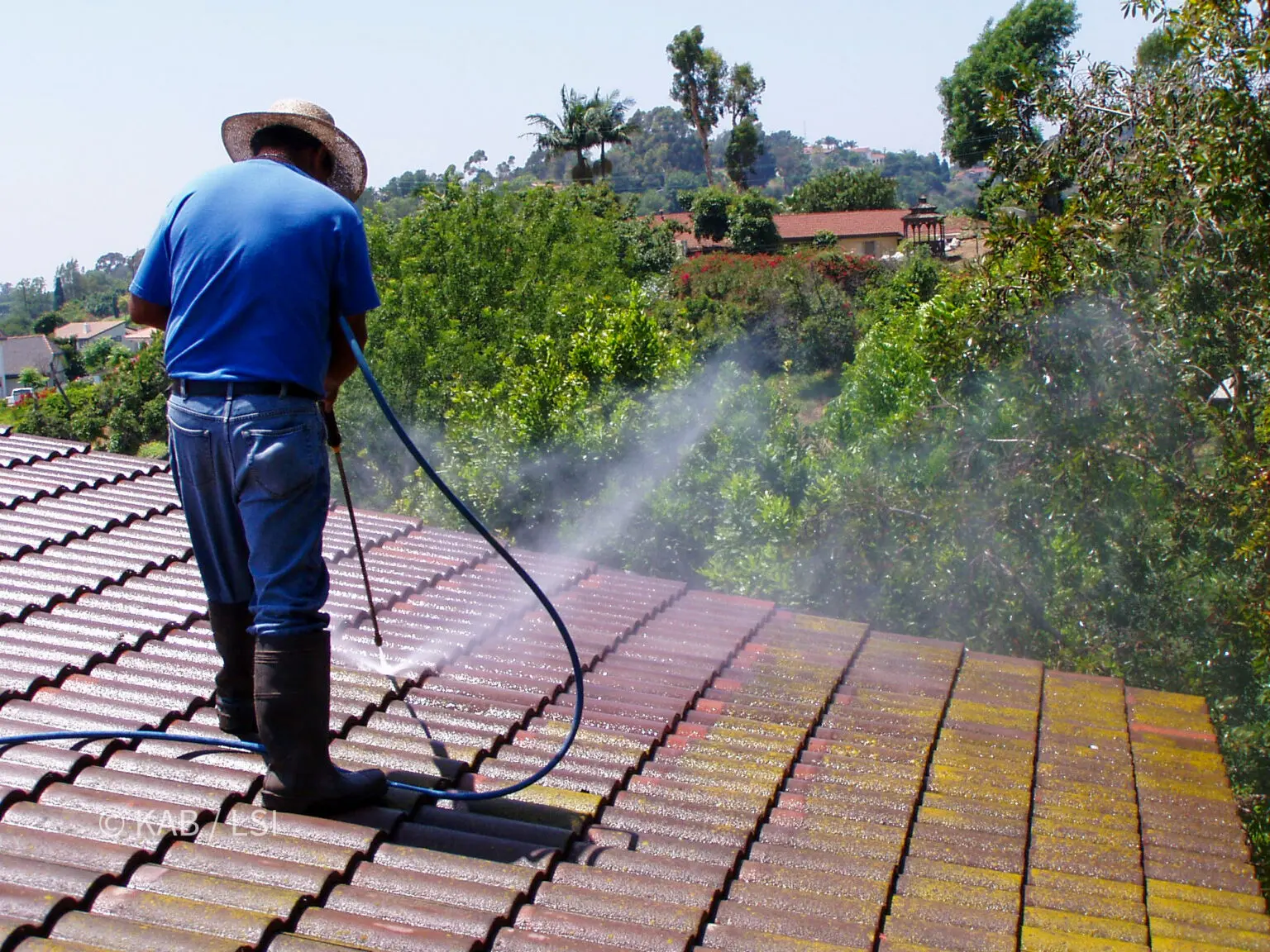 Roofer pressure washing a concrete tile roof on a hillside property with a valley view behind
