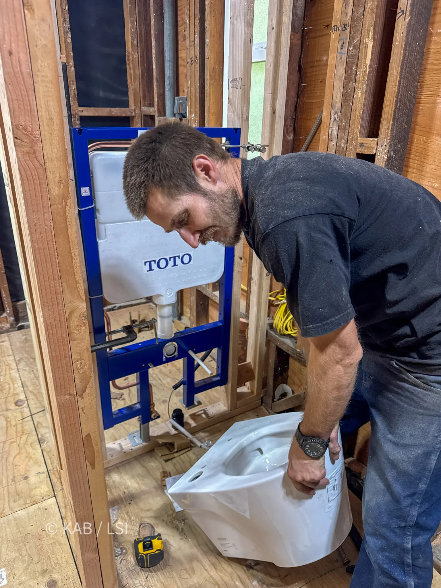 Plumber setting an in-wall carrier and wall-hung toilet into open stud framing during a bathroom remodel
