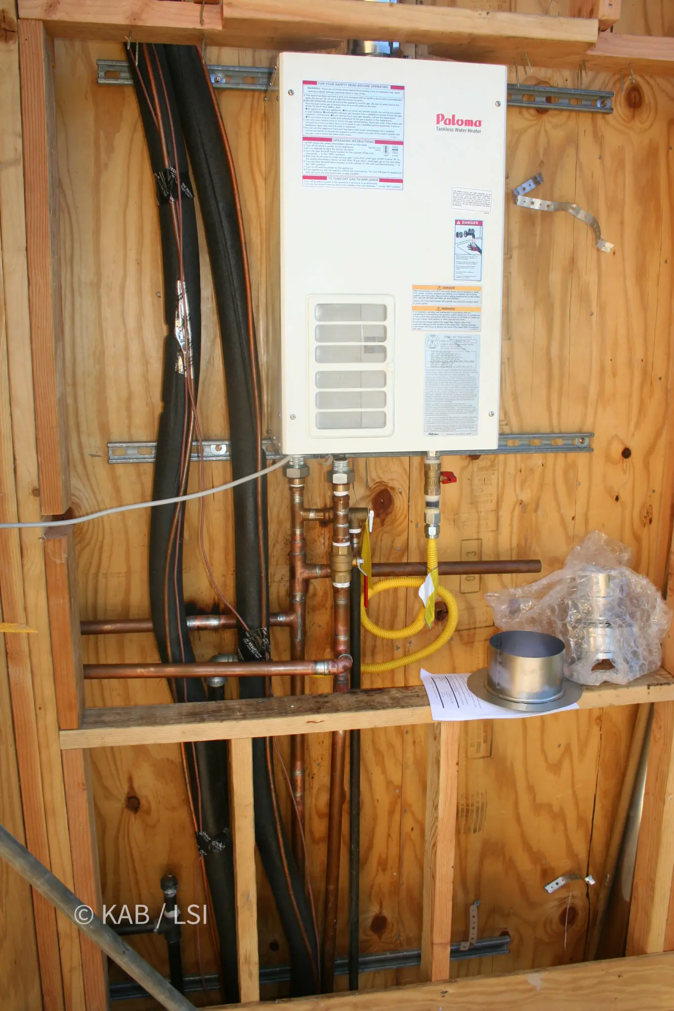 Tankless water heater mounted on open stud framing with gas line, water lines and flue clearly visible before the wall is closed