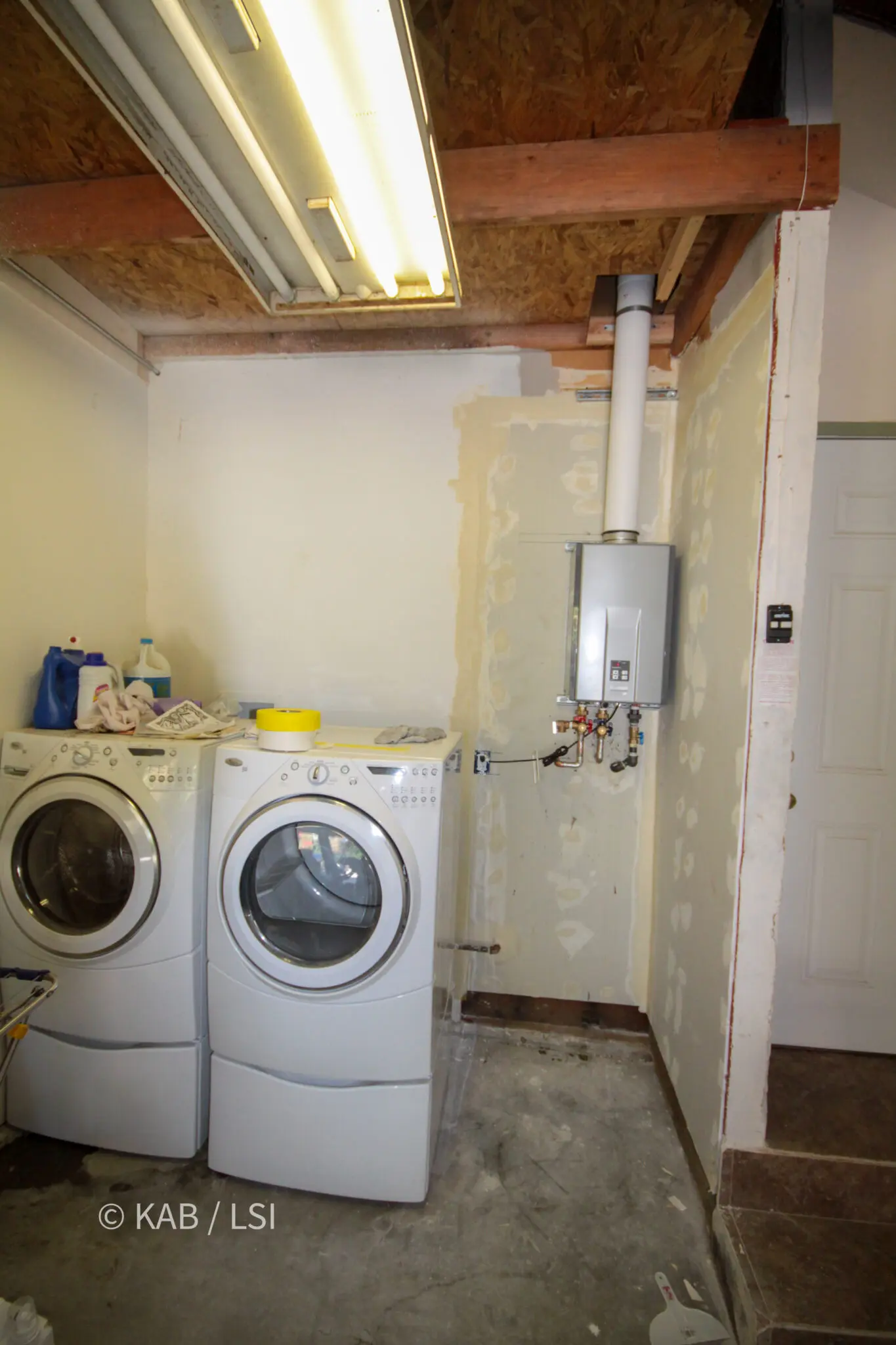 Laundry room after removal of the water heater closet, with washer and dryer in place and a wall-mounted tankless heater occupying the corner
