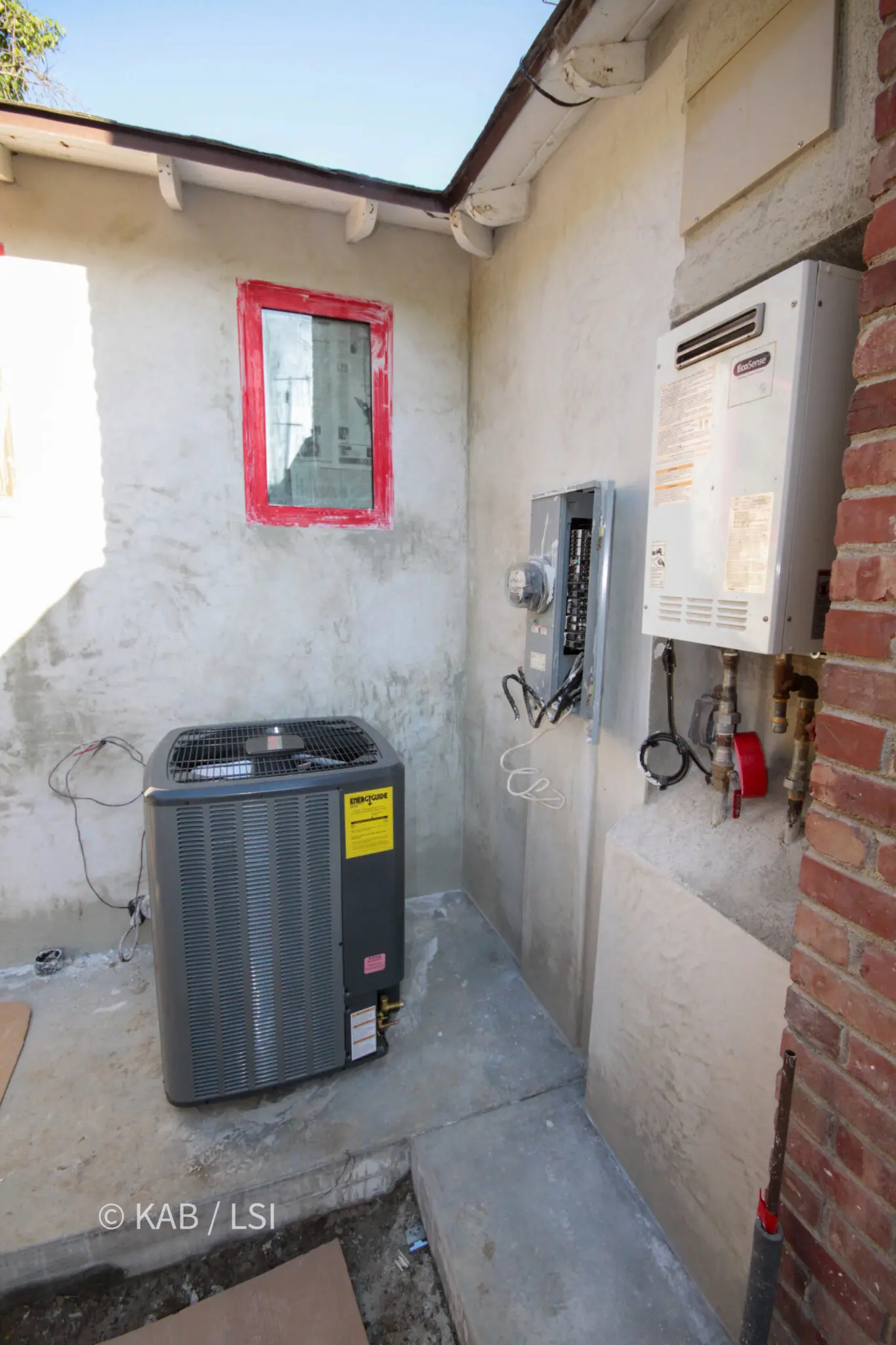 Completed exterior mechanical wall with tankless water heater, electrical service panel and HVAC condenser organized into a compact utility area