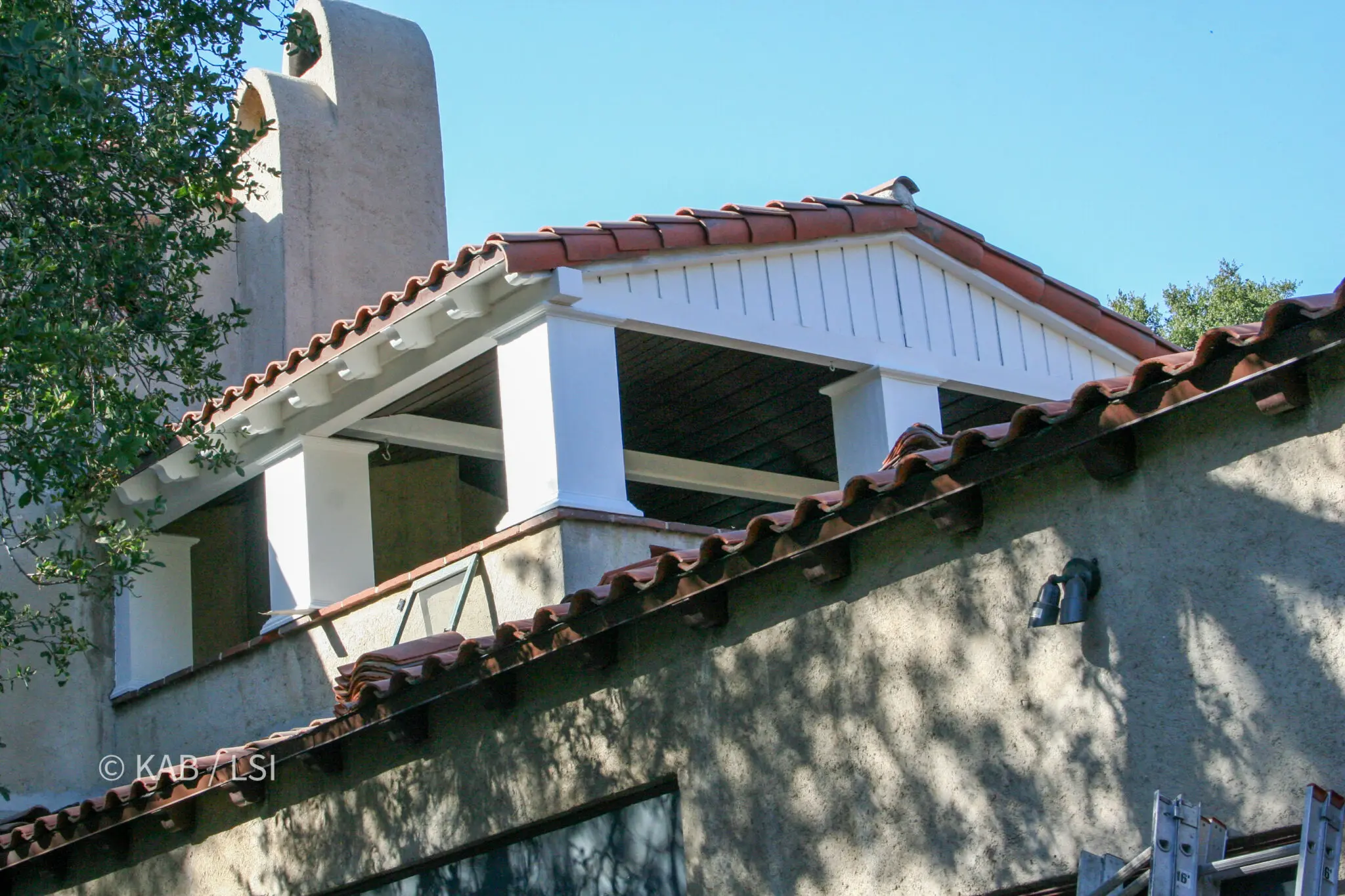 Two carpenters working on a clay tile roof, one reaching over the parapet to work on the eave below