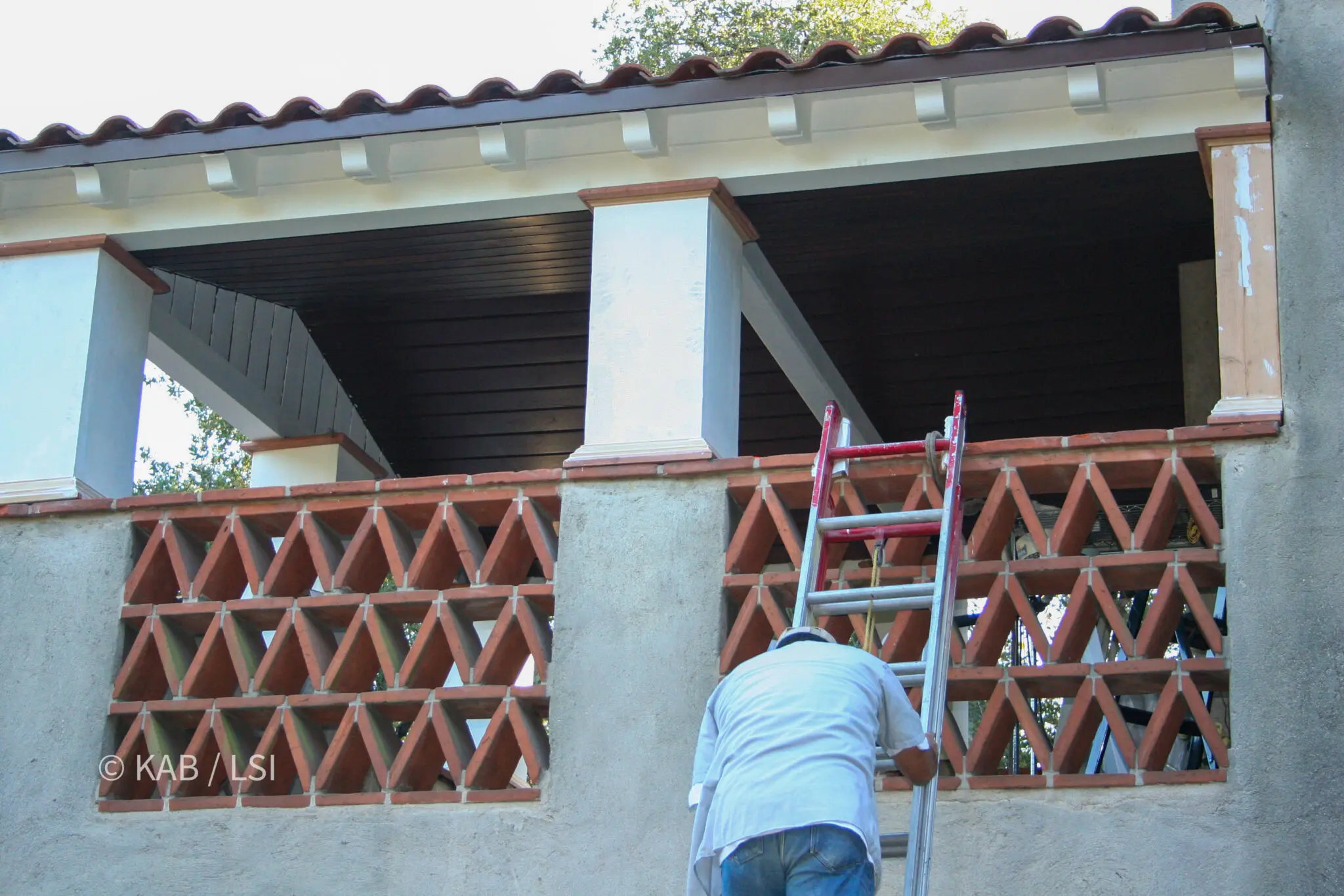 Carpenters rebuilding rotted rafter tails on the second-story loggia of a 1920s house, with a miter saw and lumber set up on the lawn below