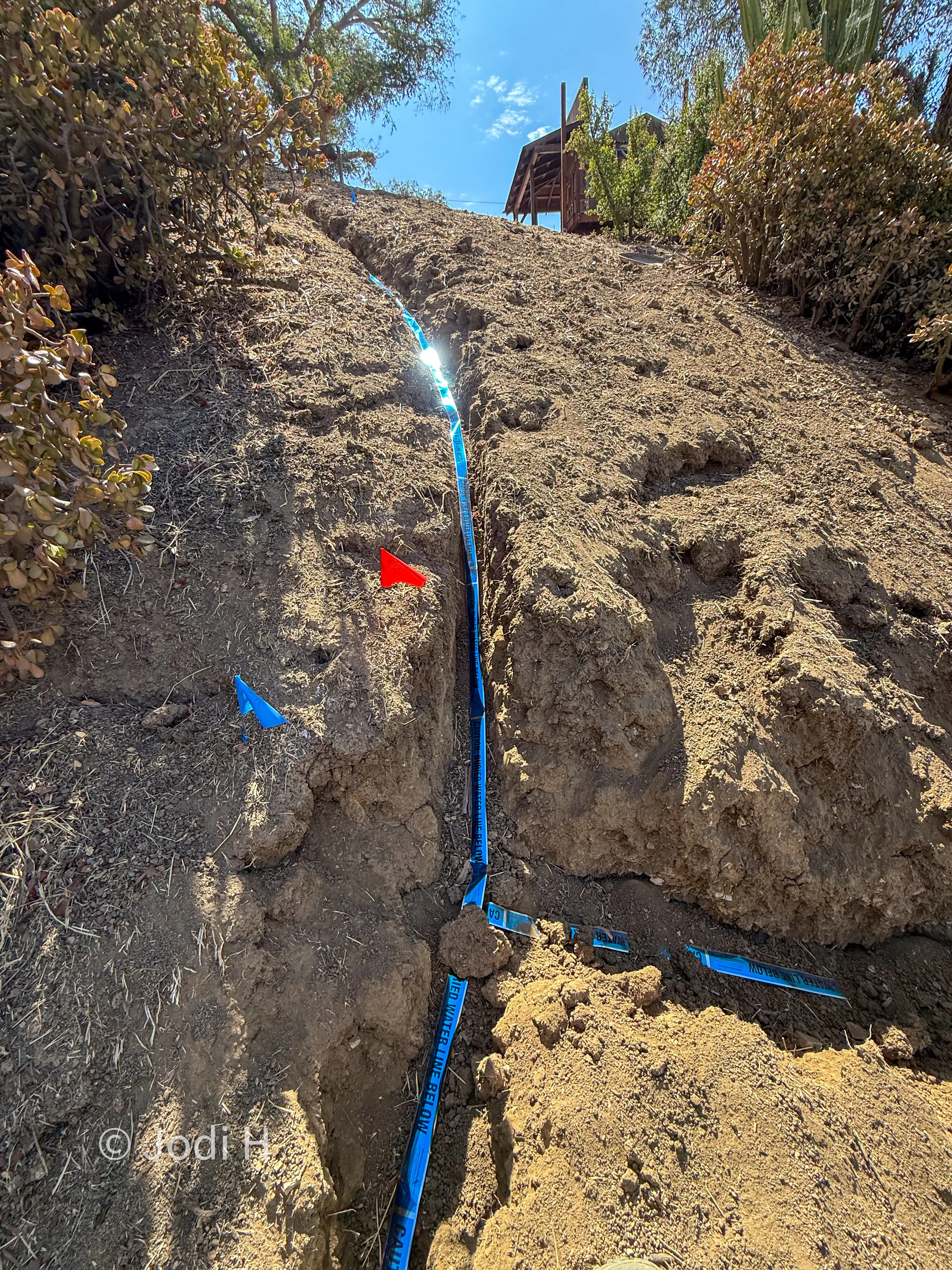 Survey flag planted in the ground beside an open water service trench to mark a buried fitting location
